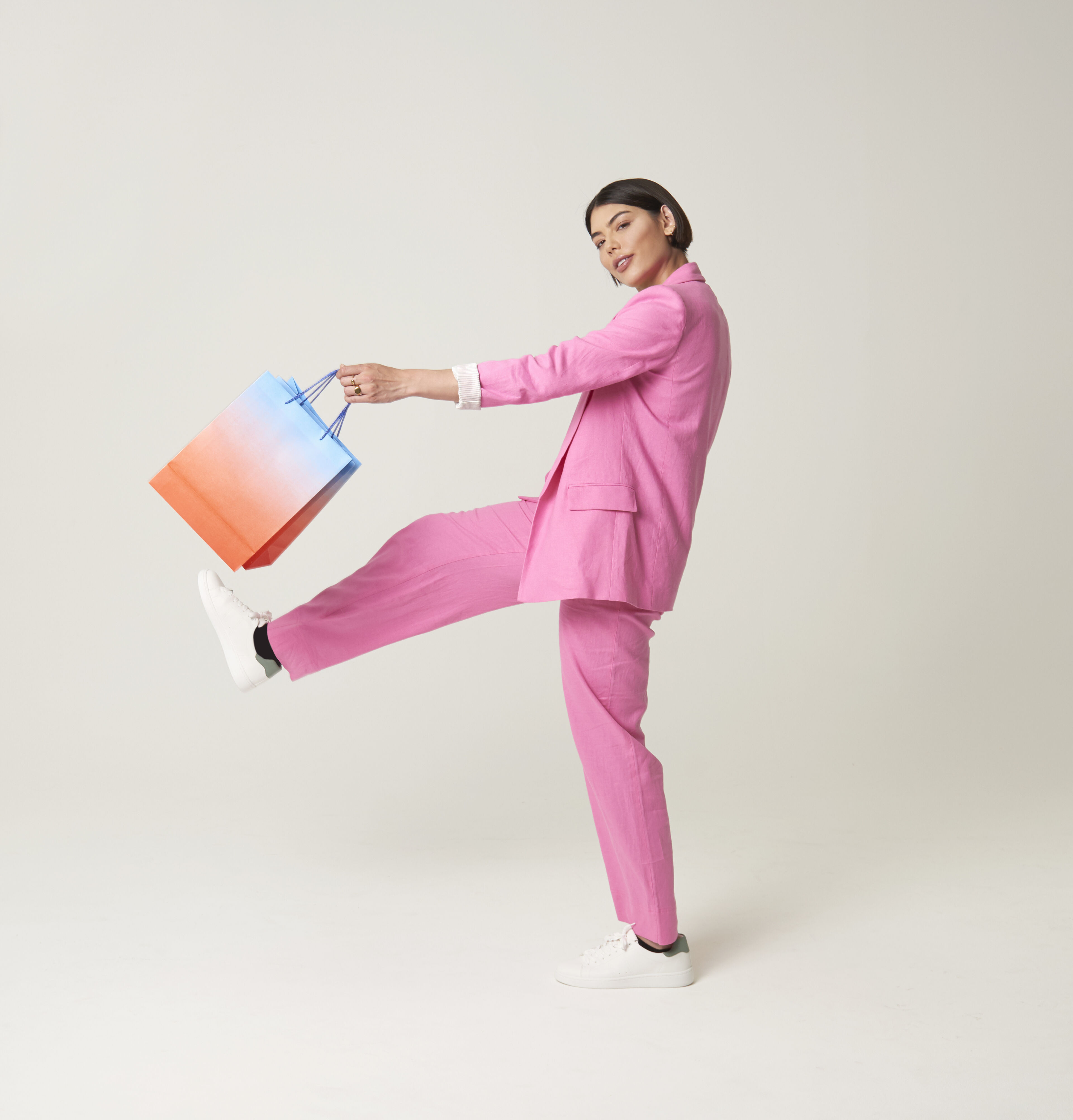 A stylish woman in a vibrant pink suit kicks up her leg while holding a colorful shopping bag, against a light background.