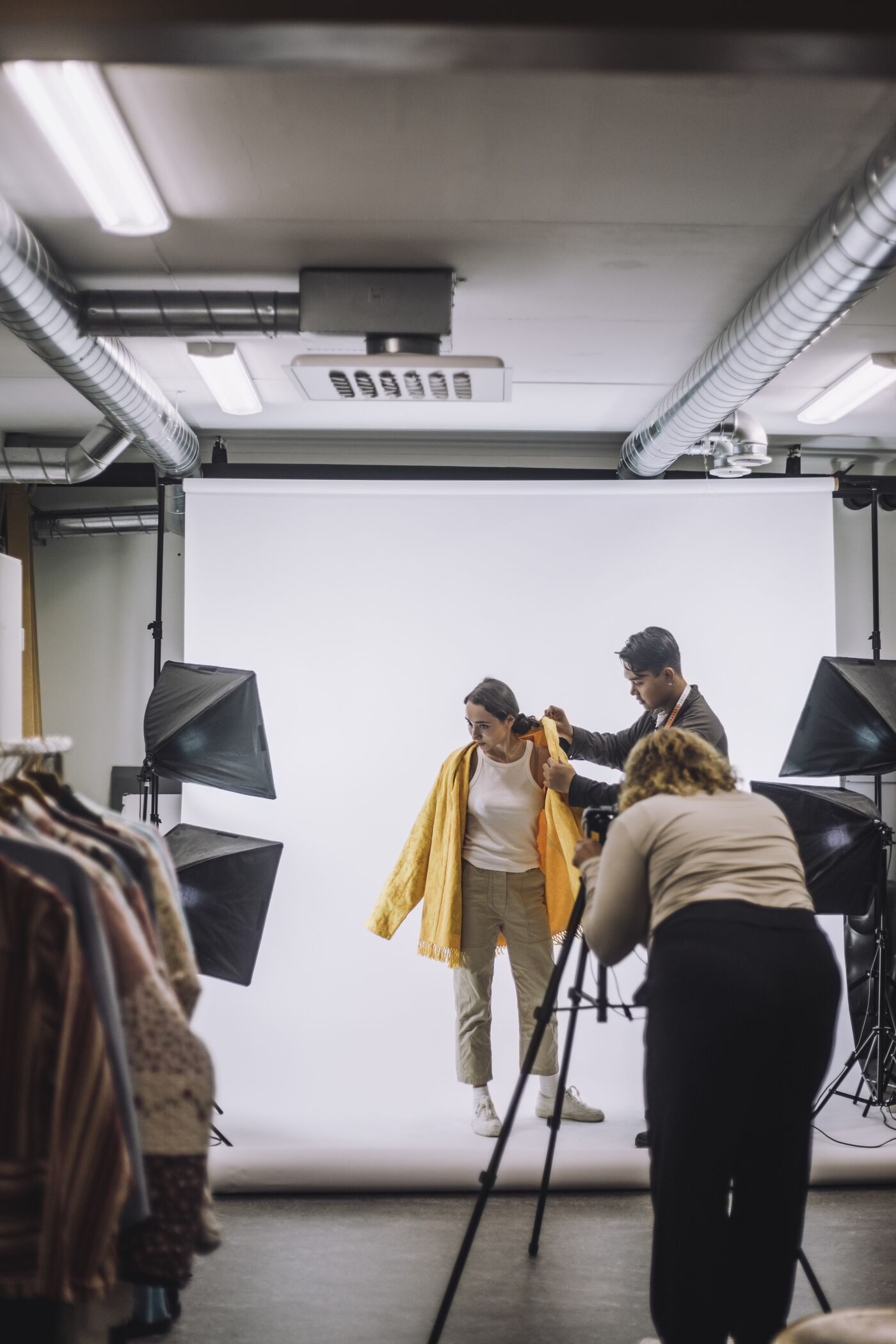 A stylist adjusts a yellow scarf on a model in a studio setup, with a photographer preparing to take pictures in the background.