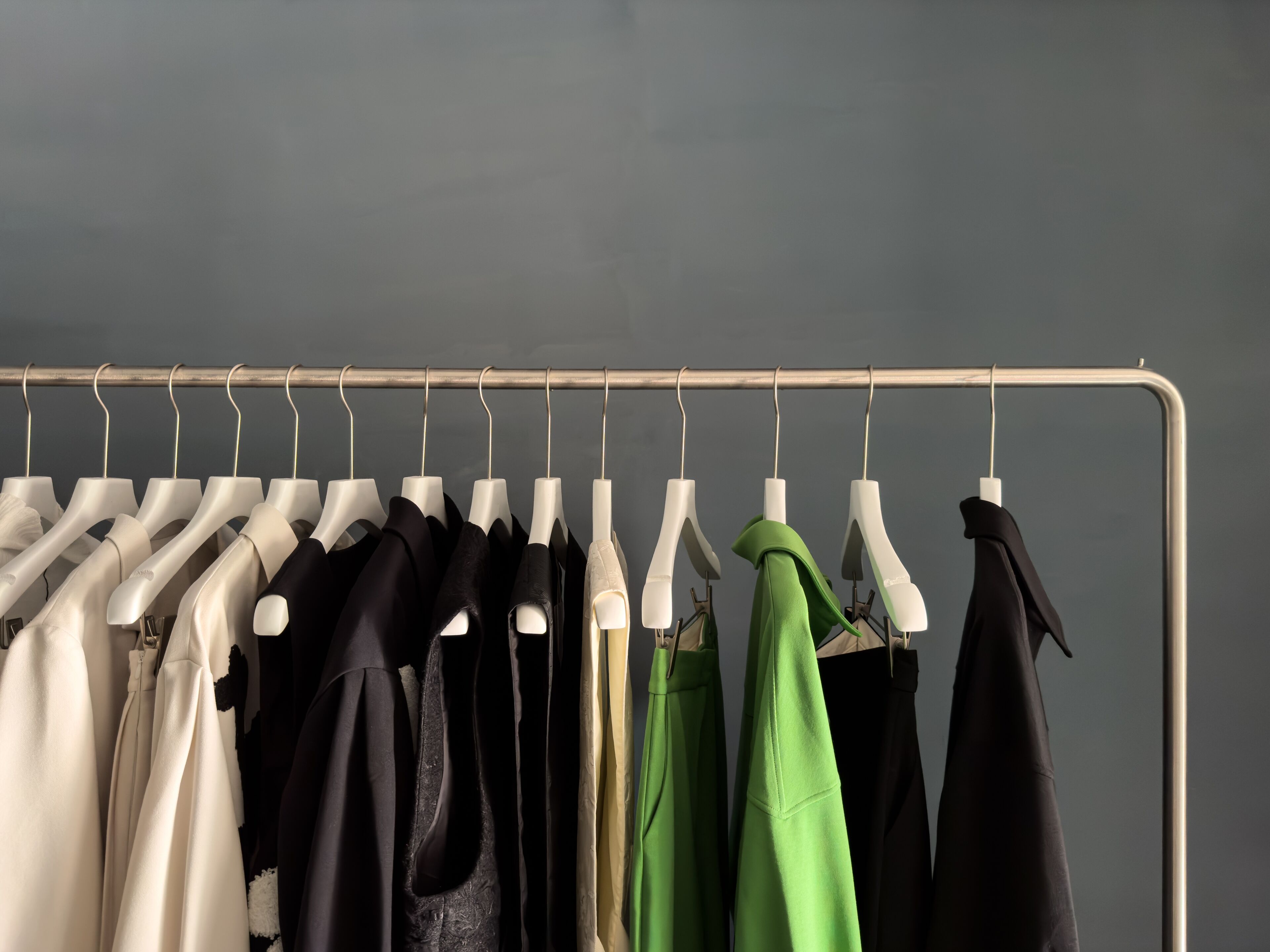 A collection of various garments in neutral tones with a striking green piece, displayed on a metal rack against a grey background.