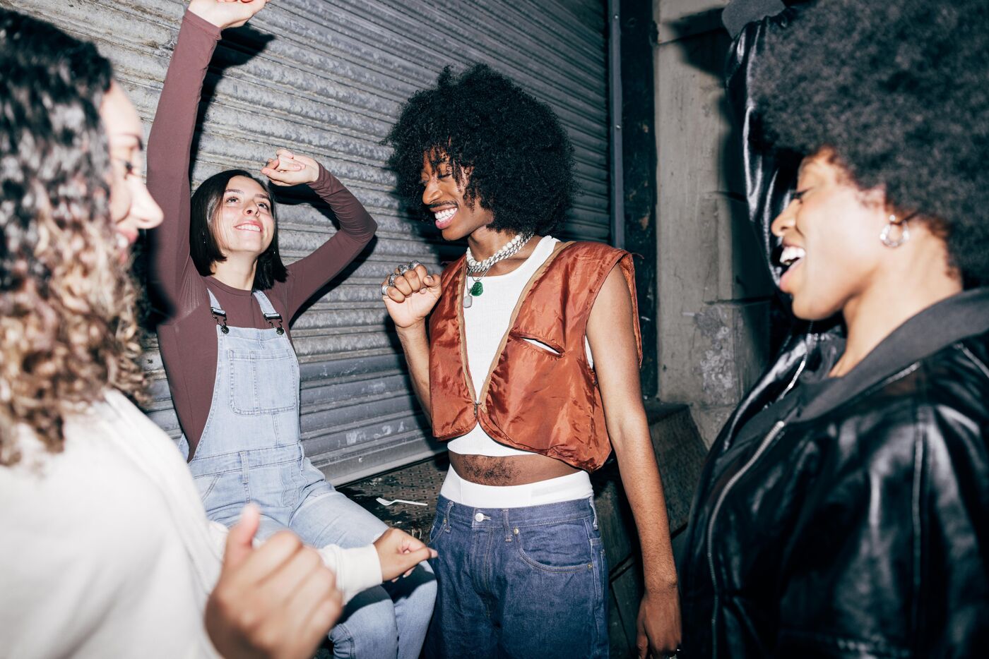 Four young women of color dance and laugh together in an alleyway at night.