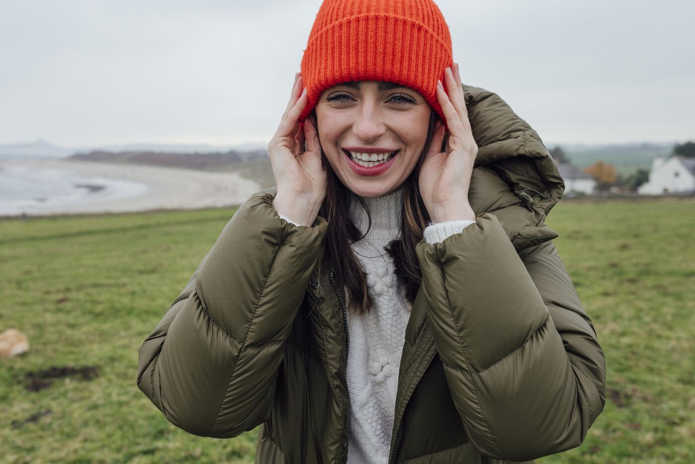 A woman wearing an orange beanie and olive green puffer jacket smiles while holding her hat on her head outdoors in a grassy field.