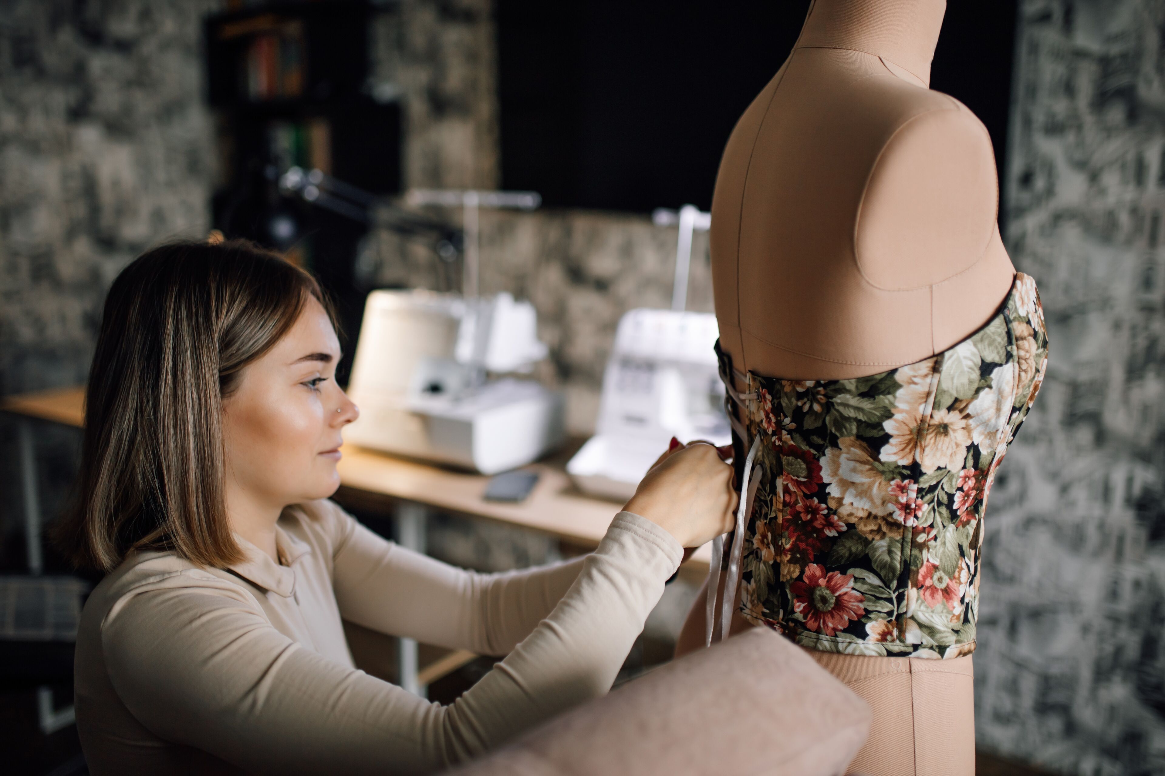 A woman tailors a floral dress on a mannequin in a design studio, suggesting meticulous handcrafting.