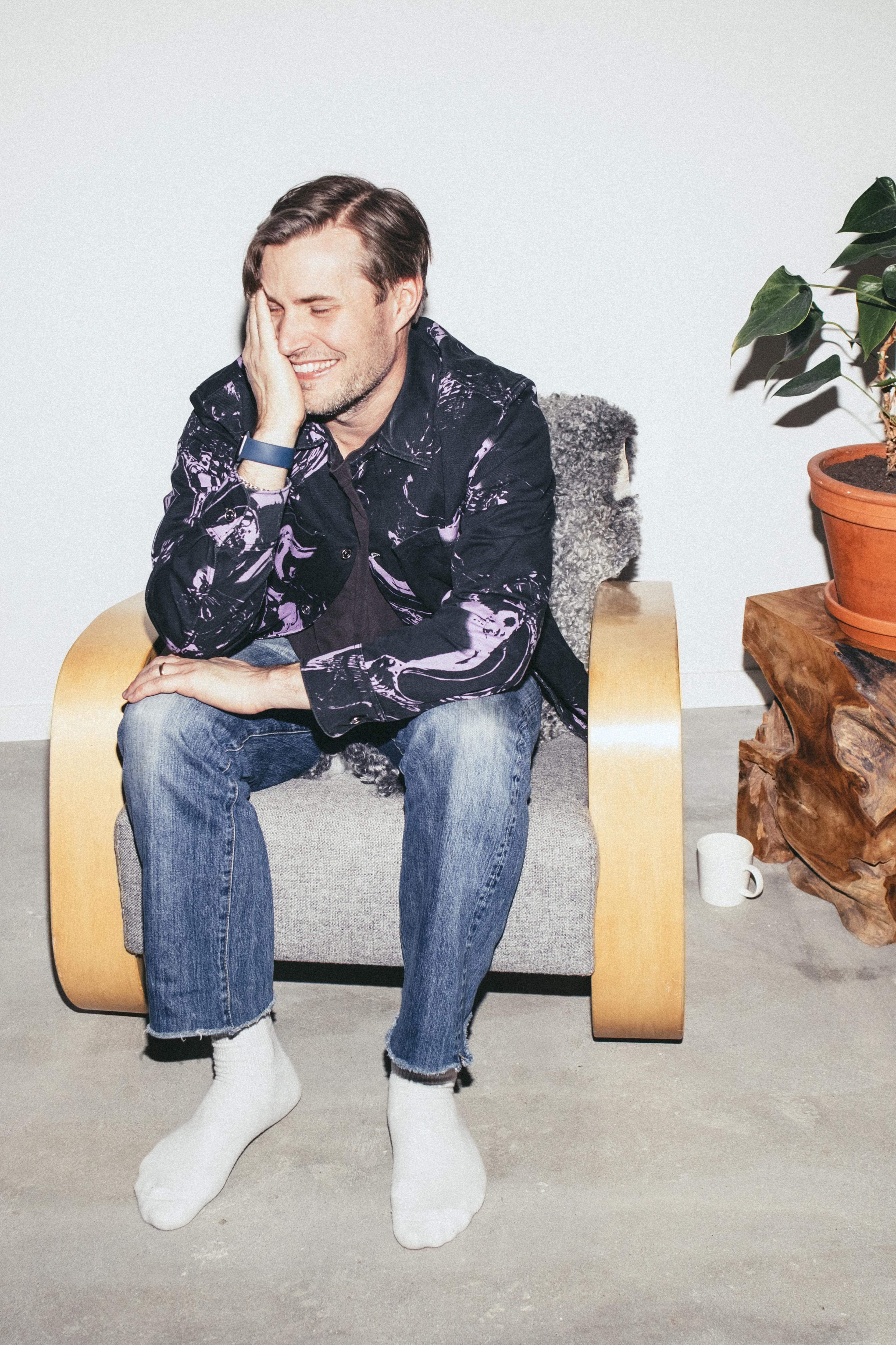 A man with short brown hair sits on a light wood chair with gray cushions, wearing a dark patterned shirt, blue jeans, and white socks, smiling while touching his face.