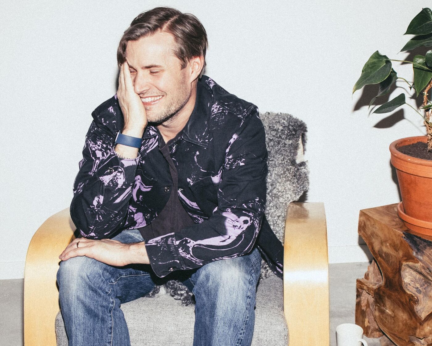 A man with short brown hair sits on a light wood chair with gray cushions, wearing a dark patterned shirt, blue jeans, and white socks, smiling while touching his face.