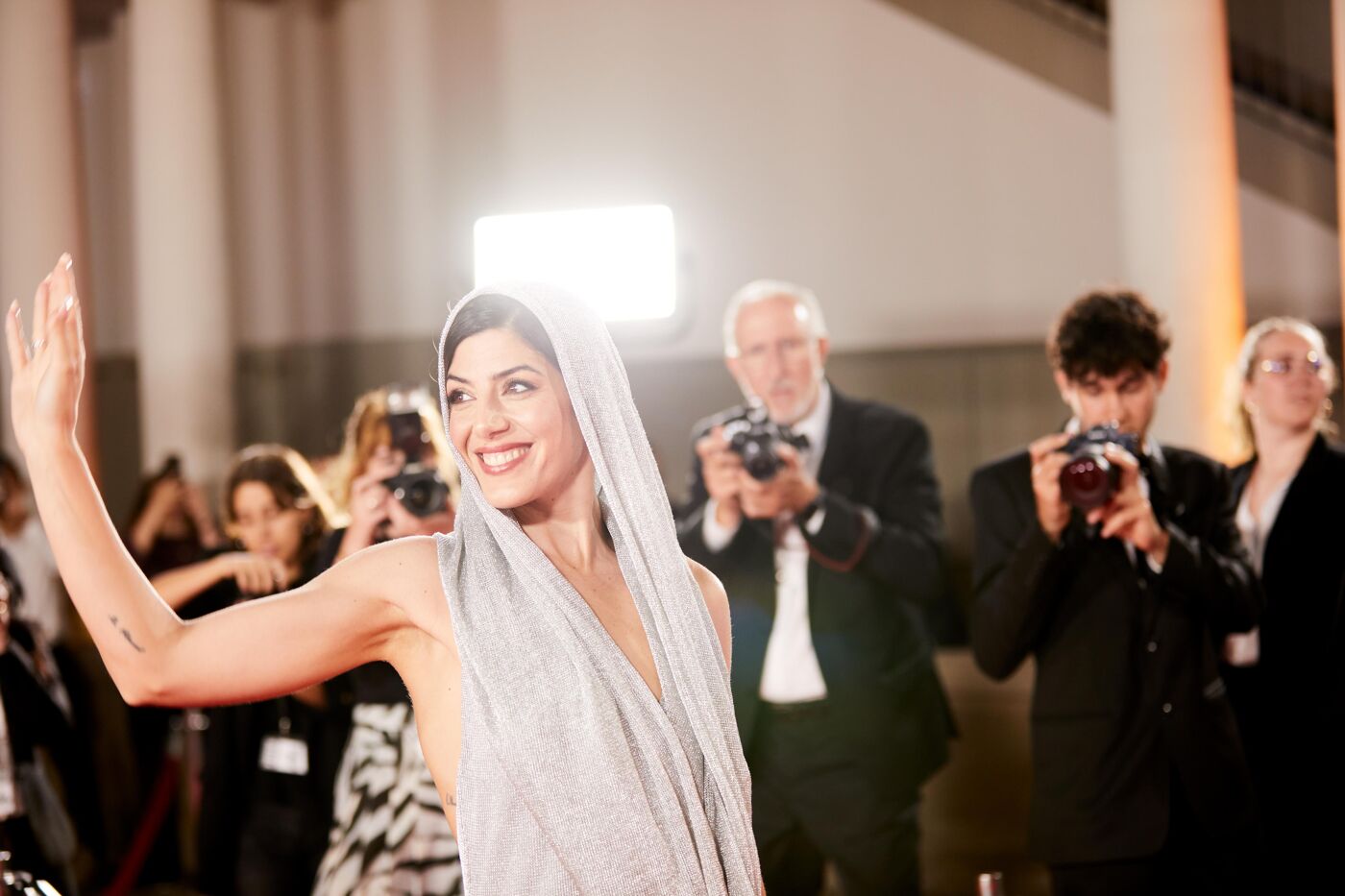 A woman in a silver hooded top waves to photographers on a red carpet.