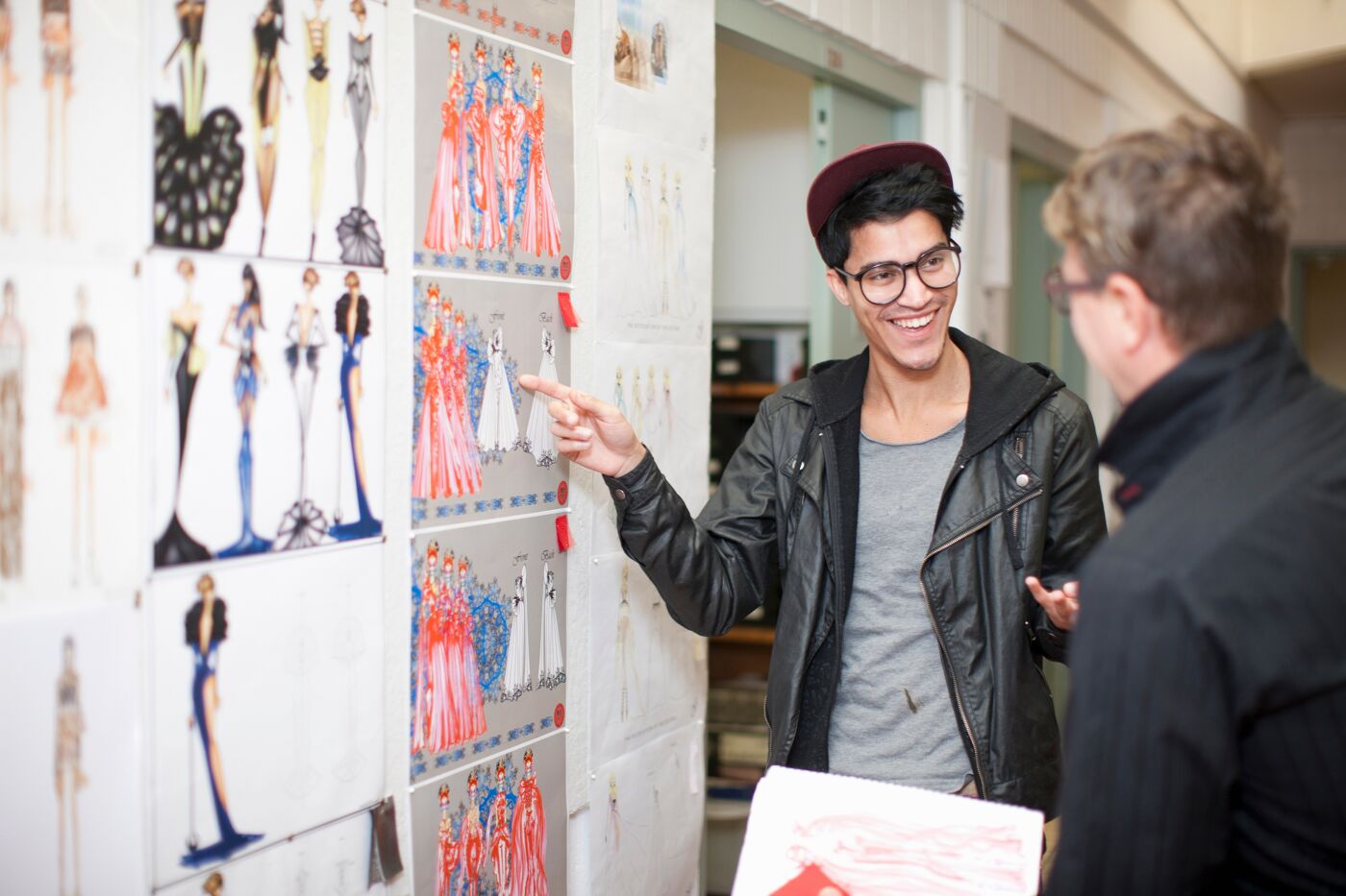 A young fashion designer discusses his colorful sketches with a colleague in a studio, showcasing a vibrant collection of evening wear designs.