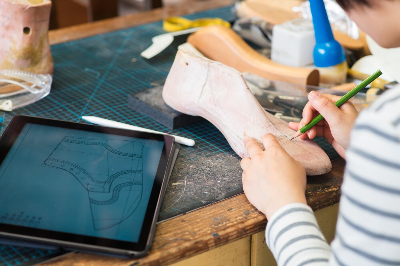 A person sketches shoe designs on a last, with a digital blueprint displayed on a tablet.