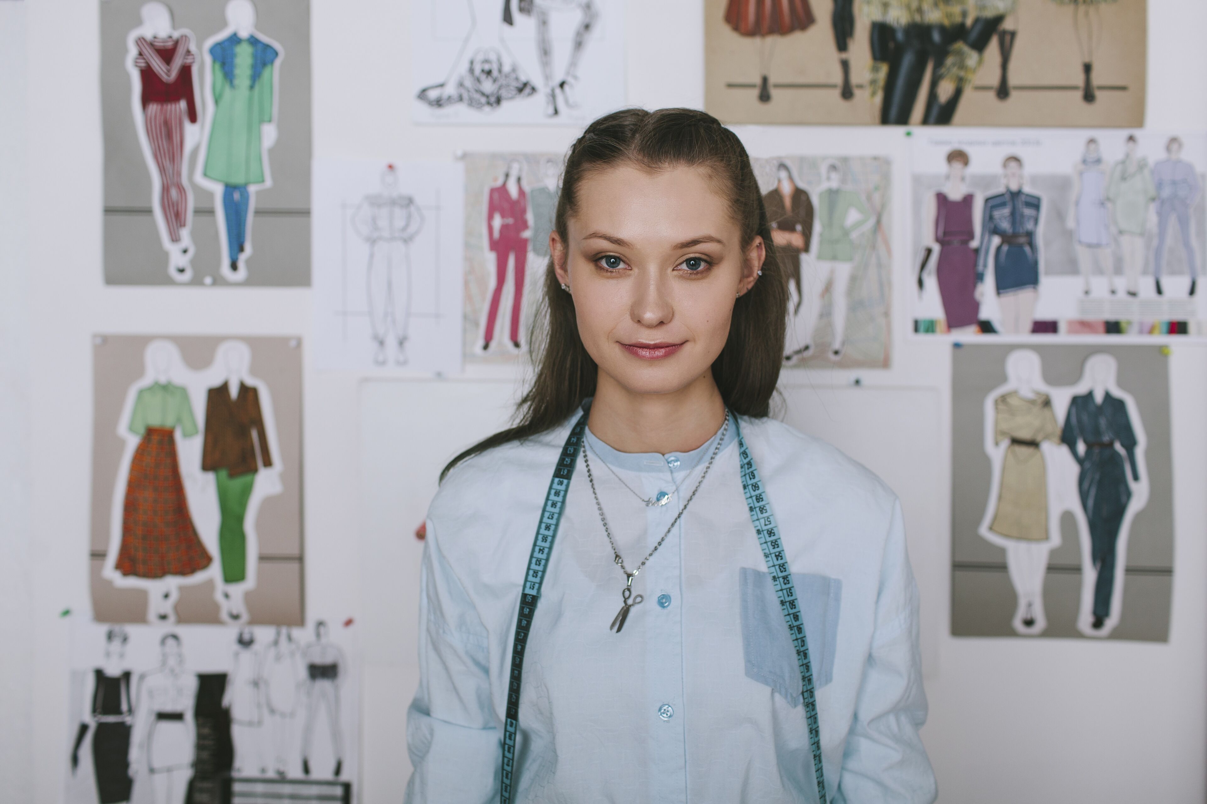 A poised young woman with a measuring tape around her neck stands in front of a fashion design mood board.