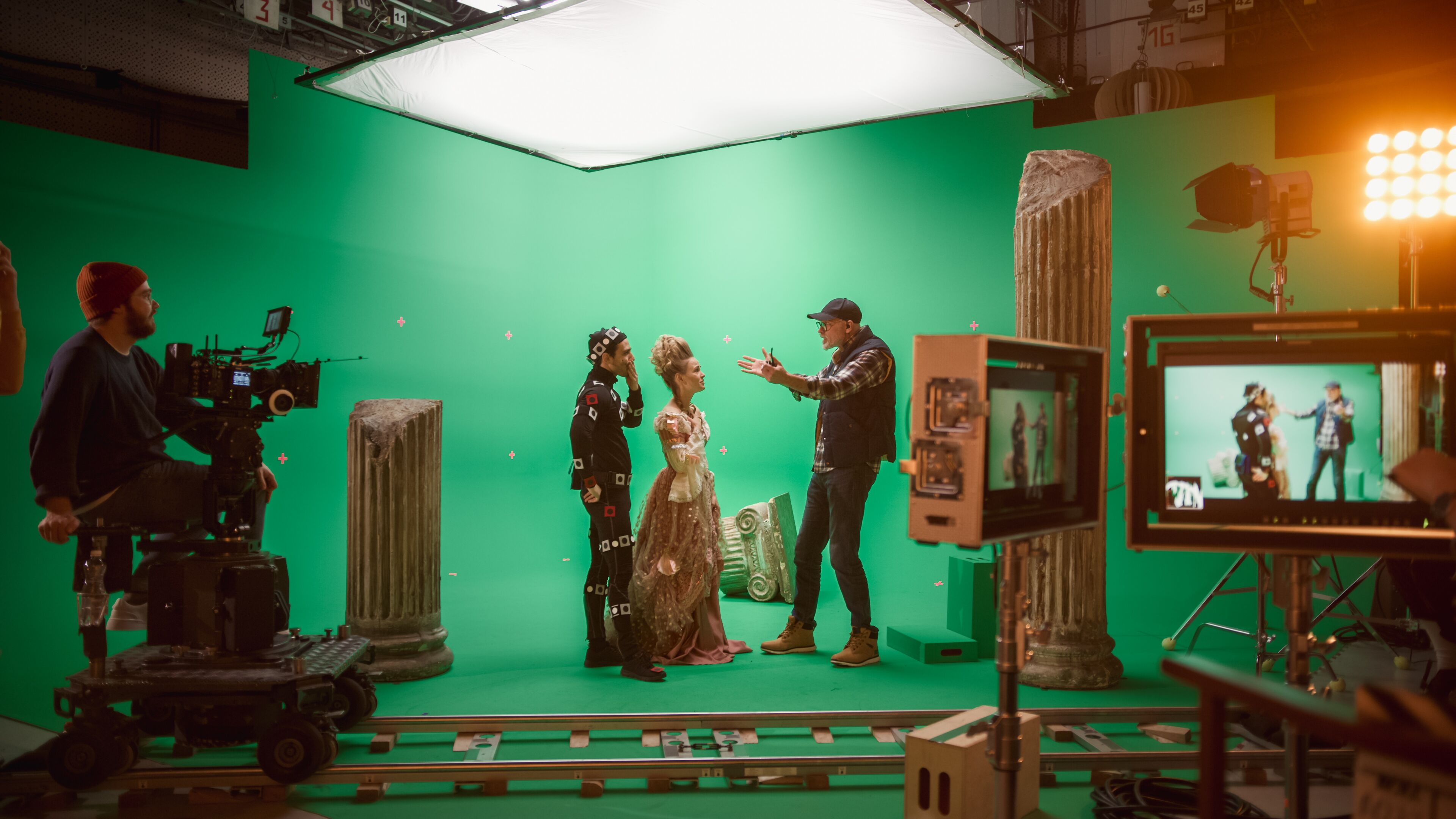 Director instructing actors in motion capture suits on a green screen set, with camera and lighting equipment.