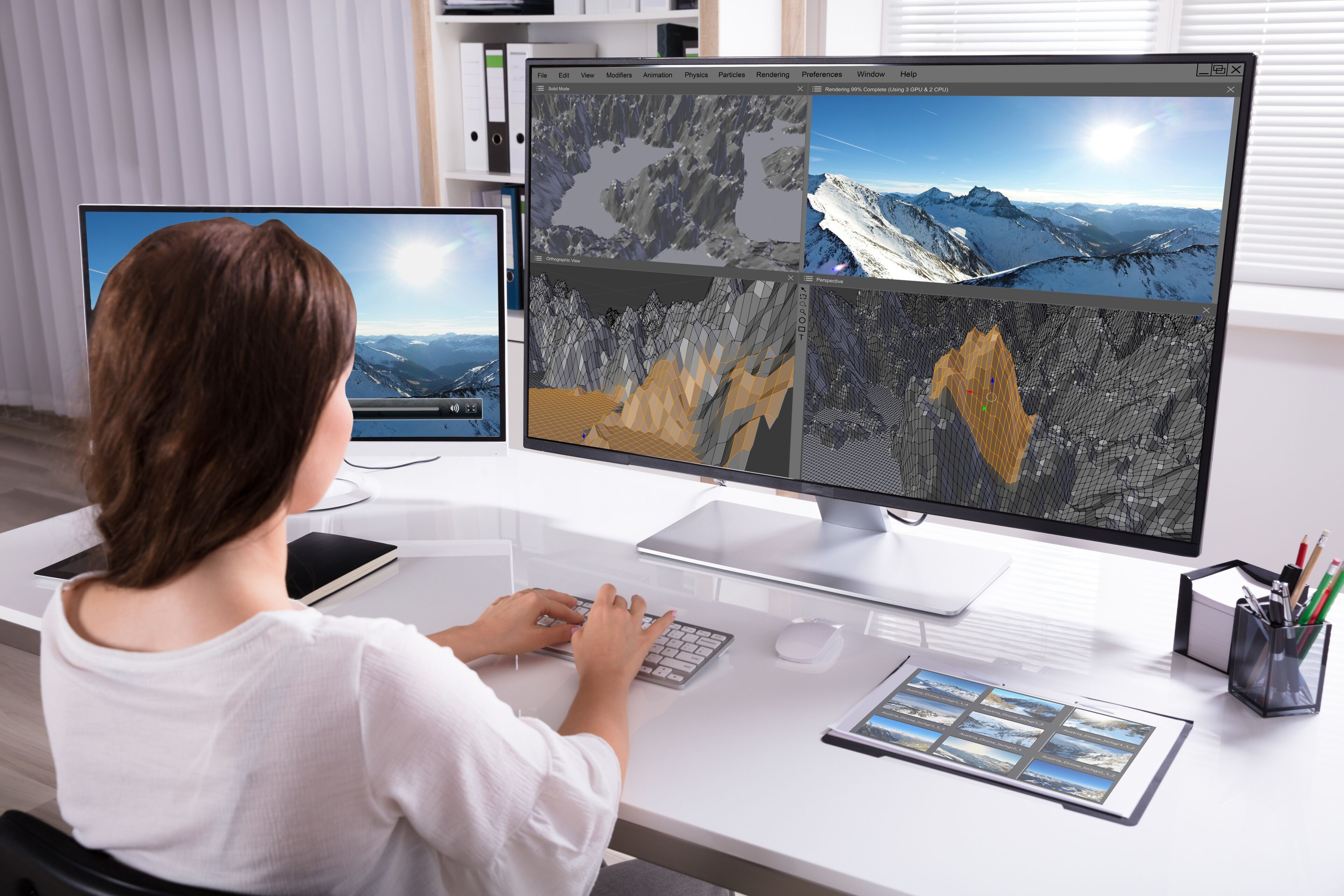 Un profesional trabaja en la renderización 3D de un paisaje montañoso en monitores dobles, con imágenes de referencia en una tableta.