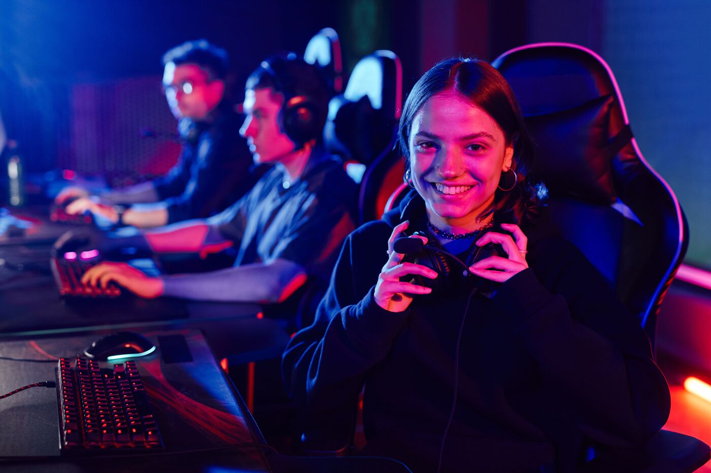 A smiling young woman with headphones around her neck at a vibrant esports gaming event, teammates in the background.