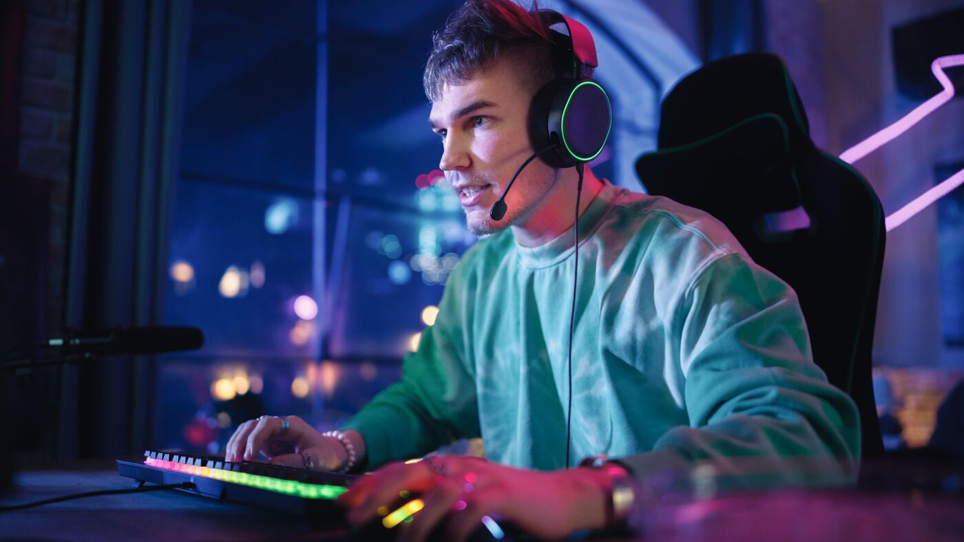 A young man wearing headphones is playing a computer game in a dark room with neon lights. He is focused on the screen, with his hands on the keyboard and mouse.