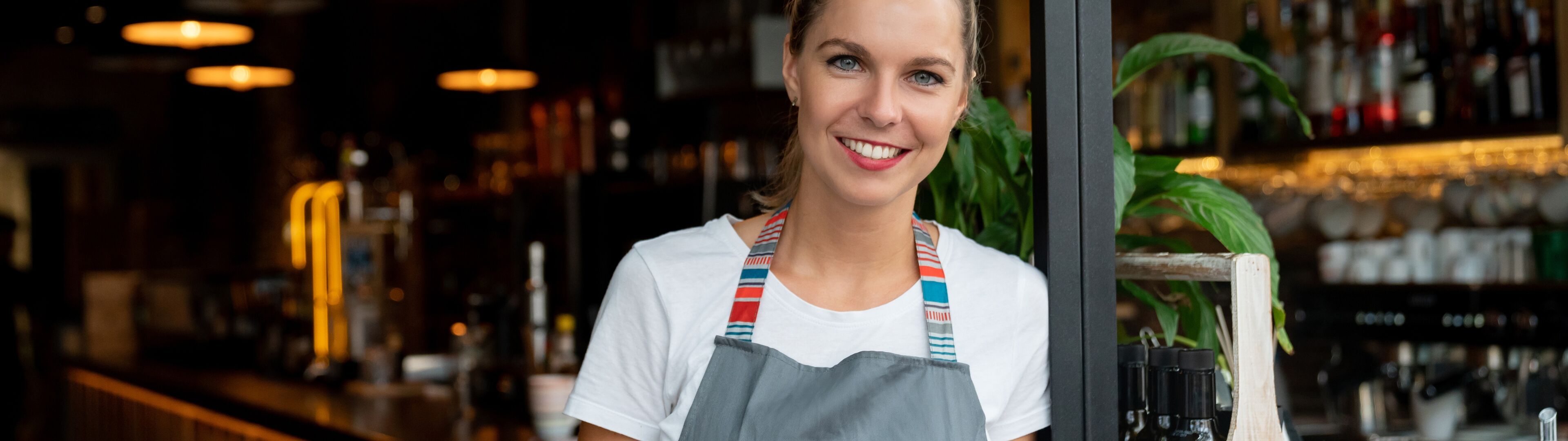 A smiling woman in a casual apron stands at the counter of a cozy restaurant, holding a digital tablet.