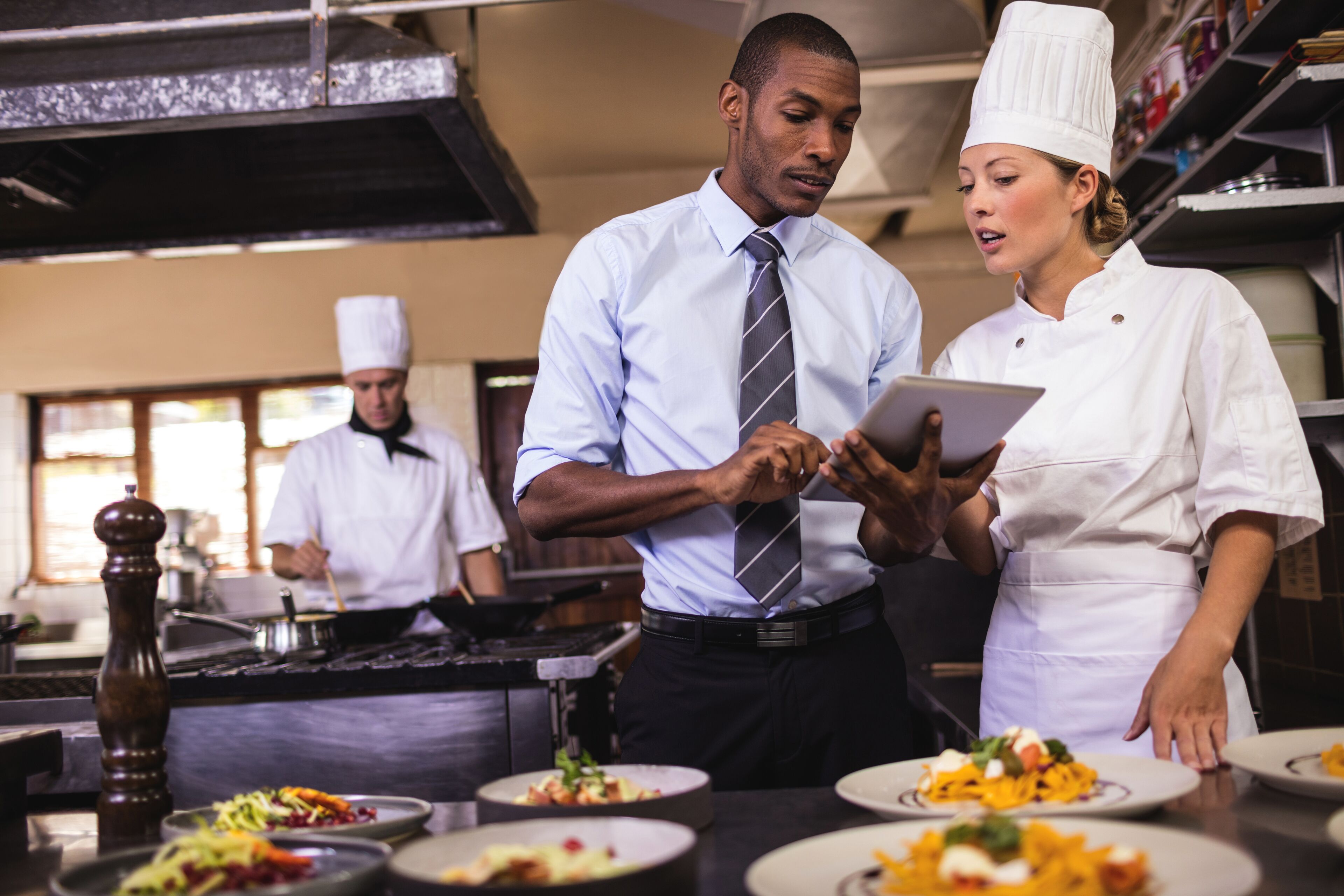 Un serveur et une cheffe discutent avec une tablette numérique dans la cuisine animée d'un restaurant, des plats prêts à être servis au premier plan.