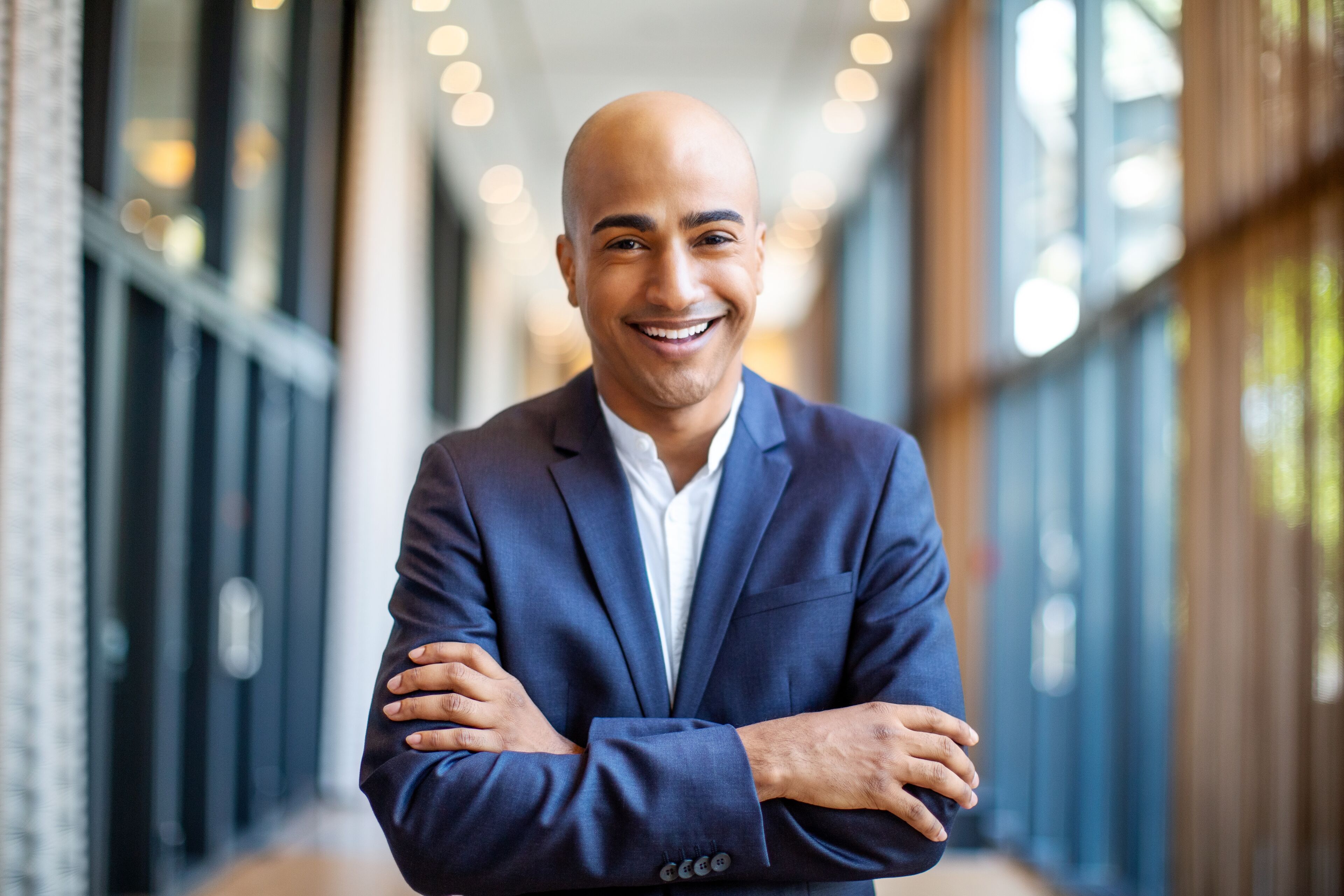 Confident bald man with a beaming smile, dressed in a navy suit, posing with arms crossed in a modern office corridor.