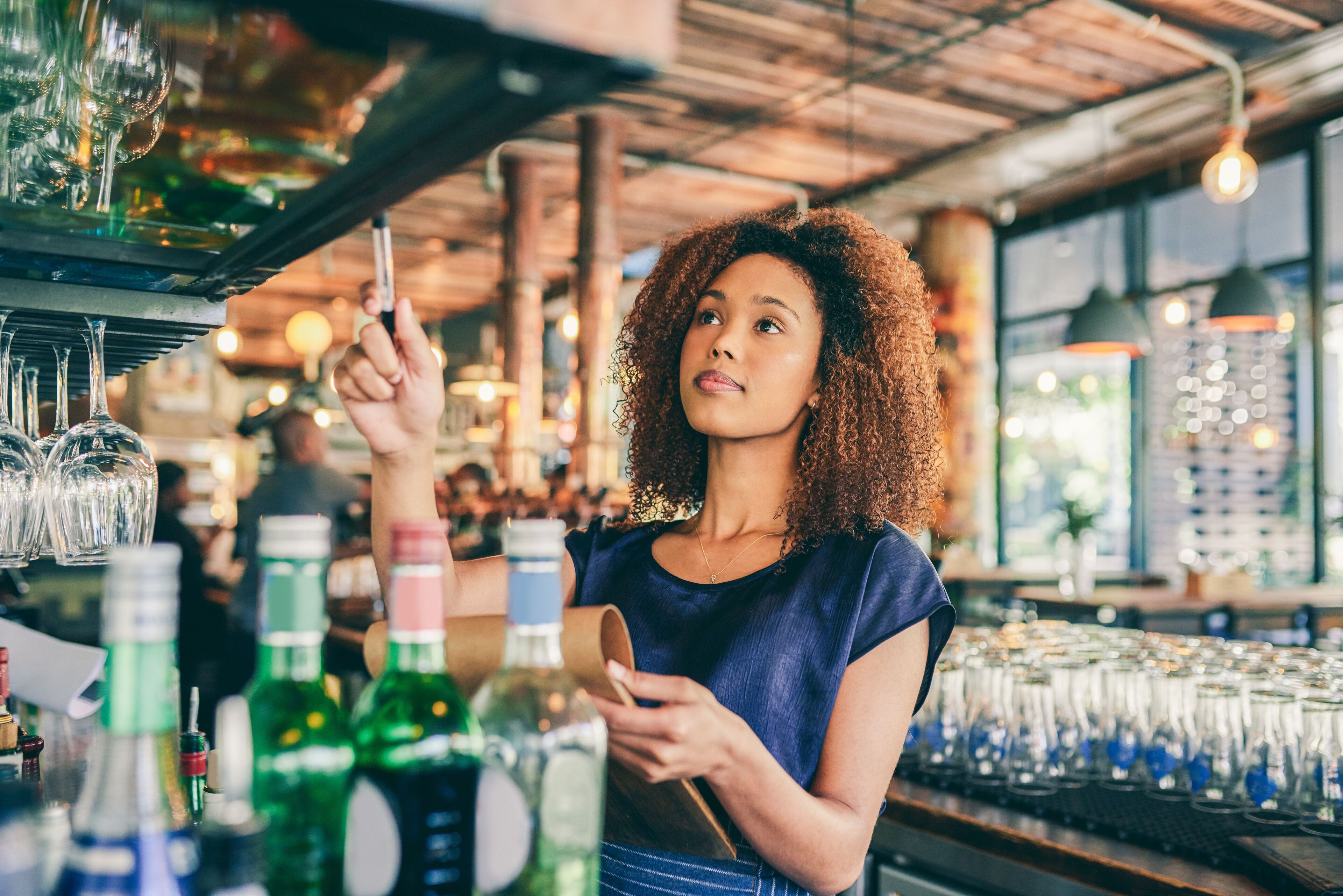 A thoughtful female bartender with curly hair is poised to take a customer's order in a bustling bar.