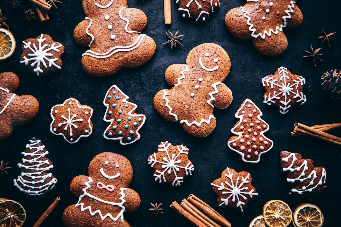 Decorated gingerbread cookies in the shapes of people, Christmas trees, and snowflakes are artfully arranged on a dark surface, accented with cinnamon sticks and dried orange slices.
