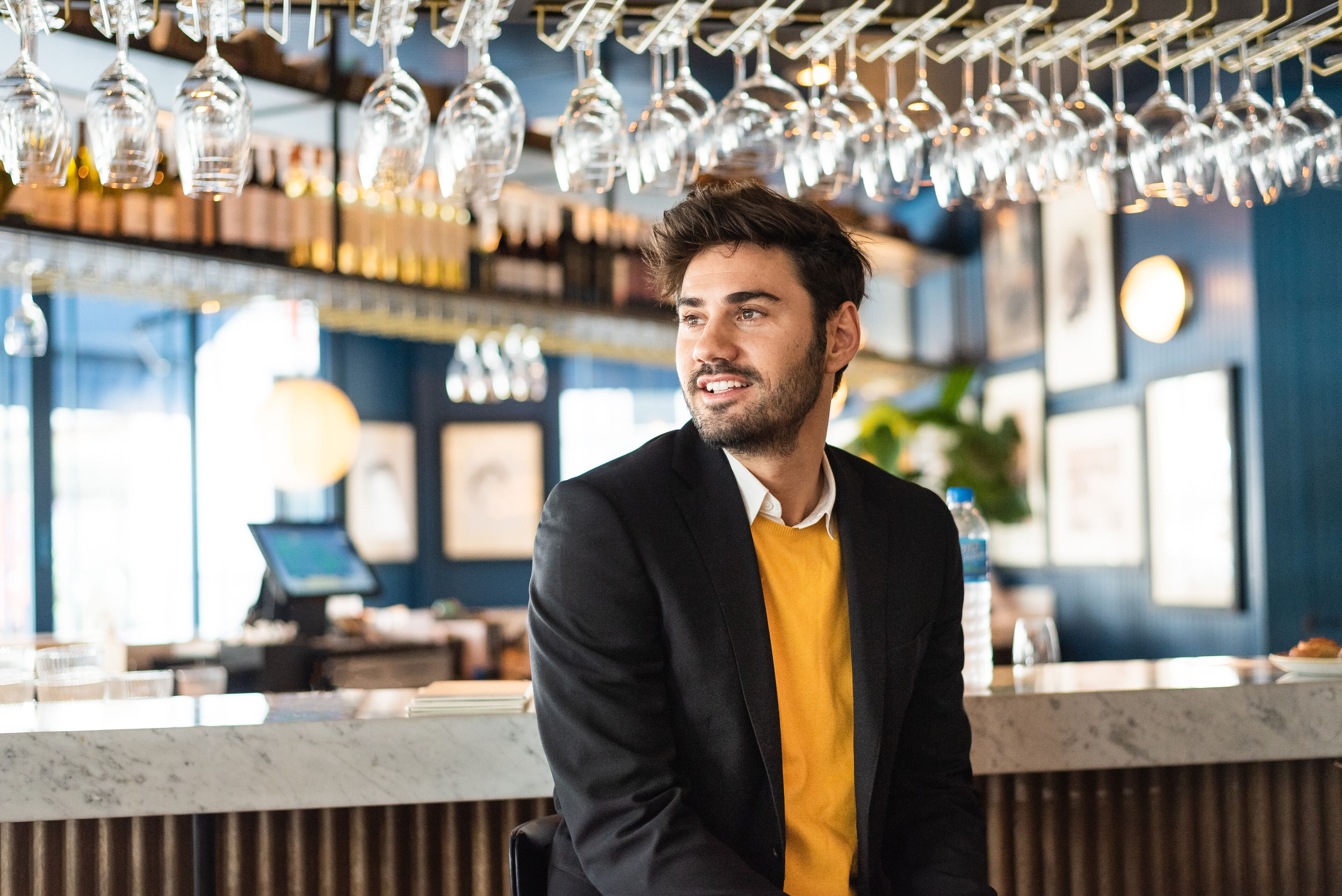 A man in a smart-casual attire at a bar, radiating confidence with a subtle smile.