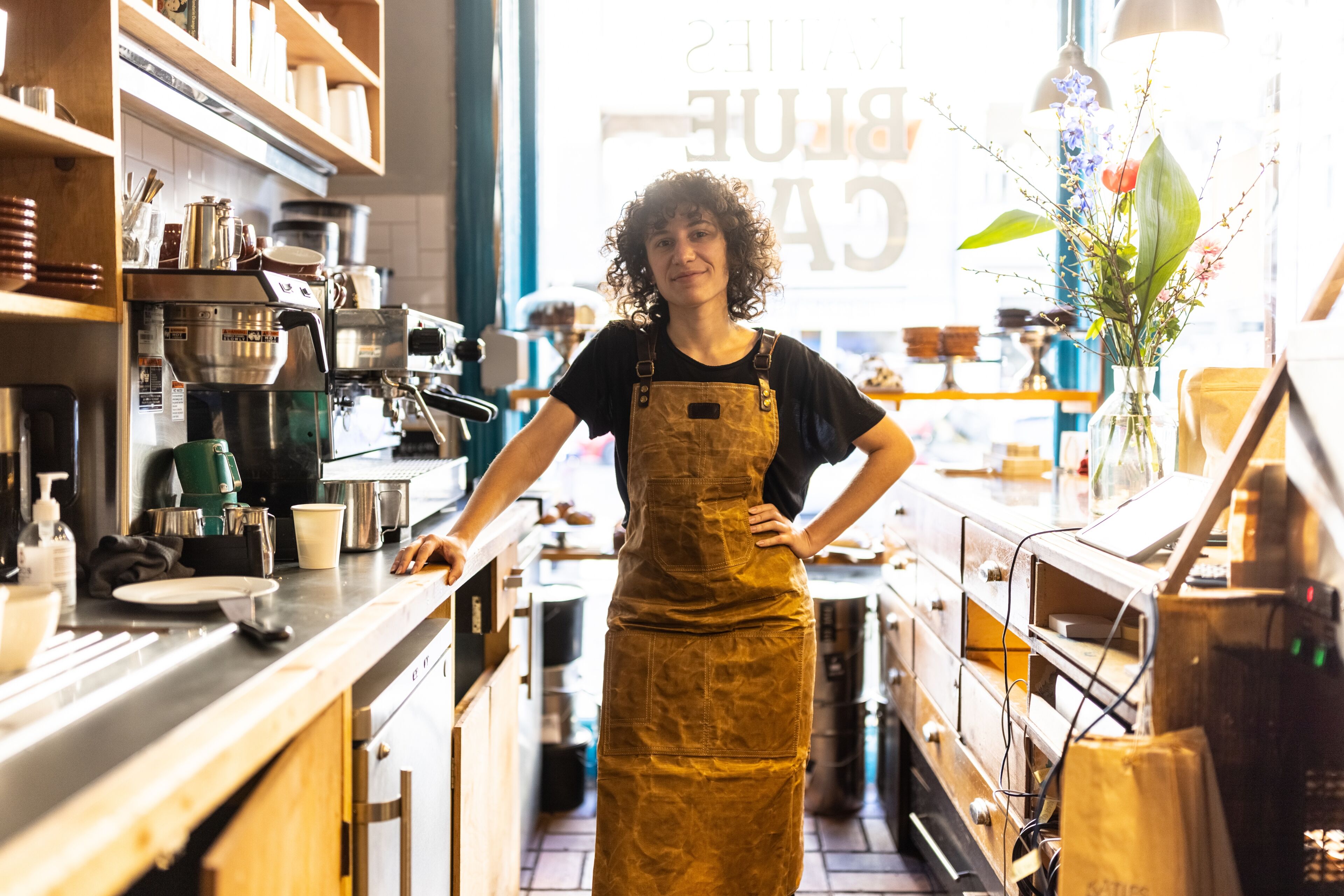 A confident female barista wearing a brown apron stands behind the counter in a well-lit coffee shop.