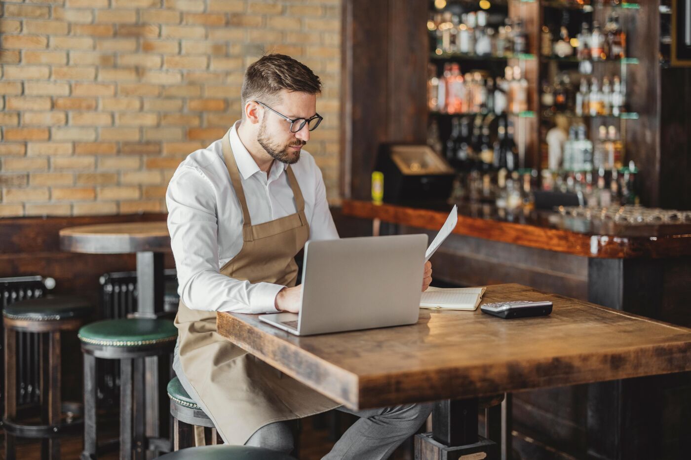 A man wearing an apron and glasses works on a laptop in a bar, with bottles in the background.