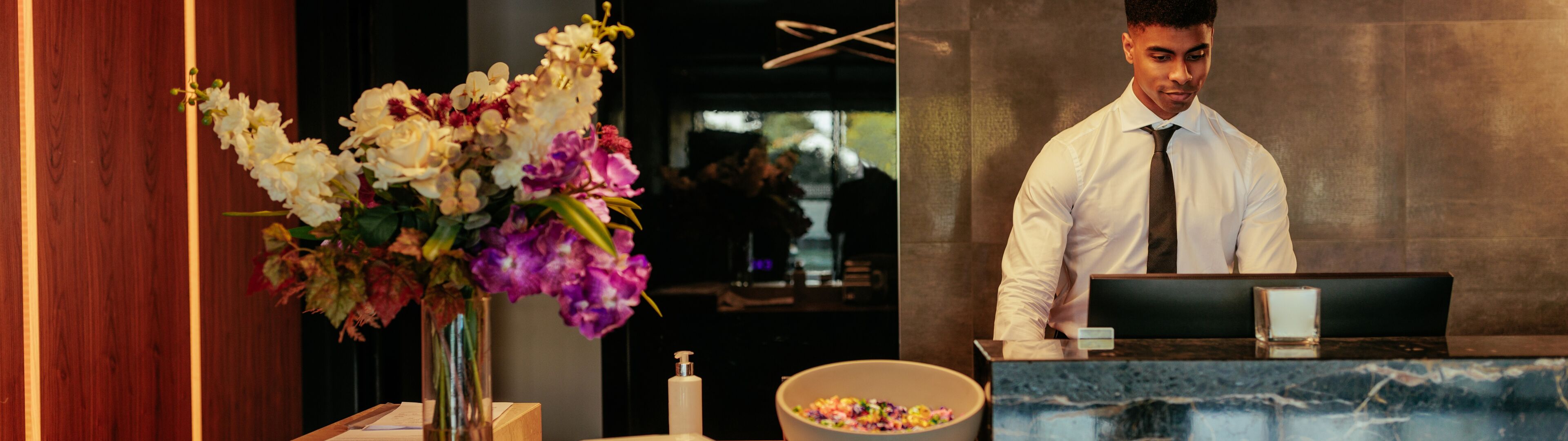 Un réceptionniste homme professionnel travaille à la réception moderne d'un hôtel, avec des arrangements floraux élégants et un éclairage stylé en arrière-plan.