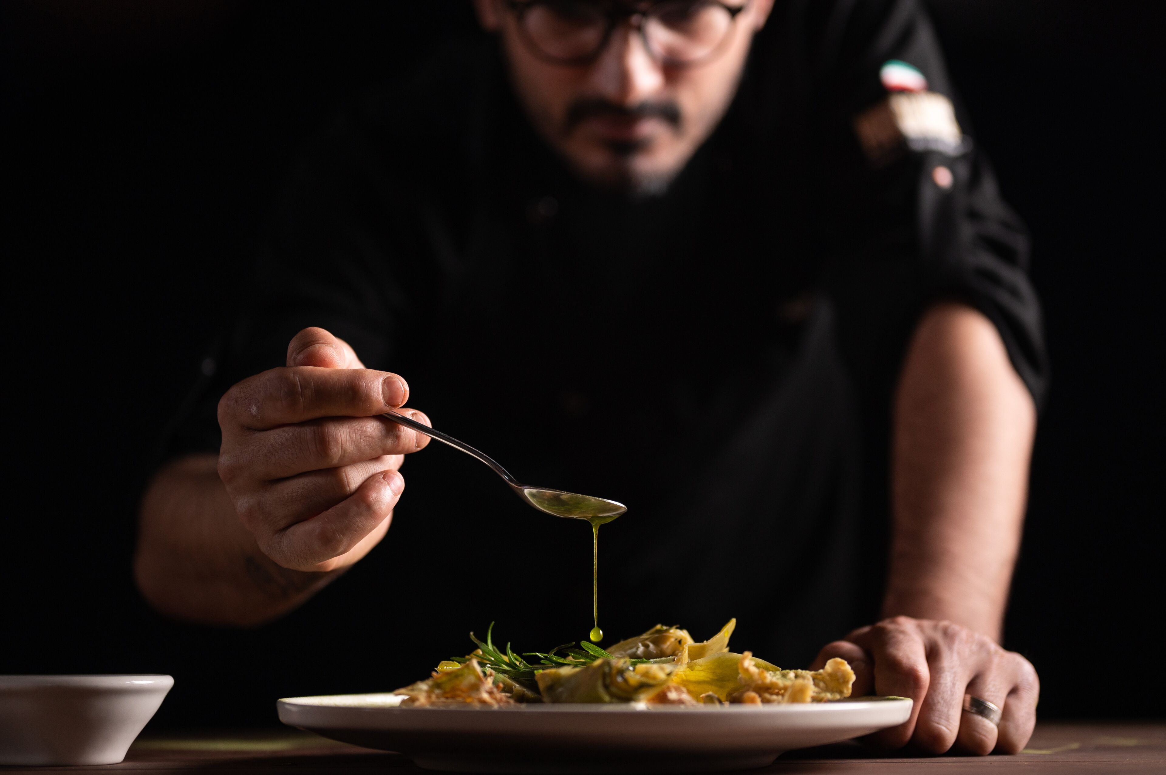 Chef enfocado añadiendo el toque final a un plato gourmet con una cuchara, en una cocina con luz tenue.