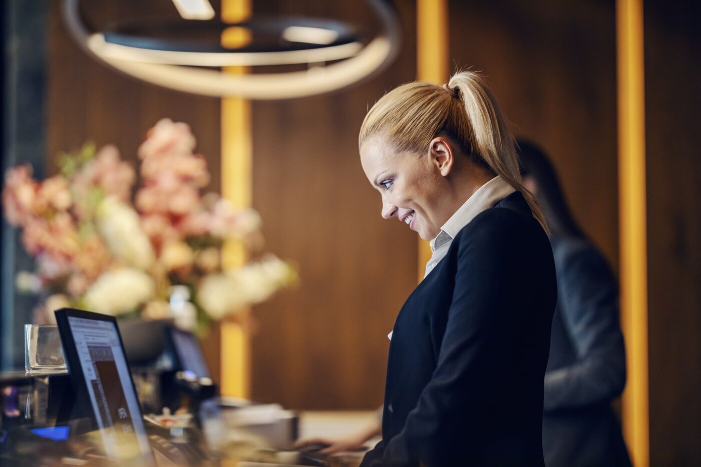 A professional female hotel receptionist smiling while using a computer at the reception desk, with a blurred background.