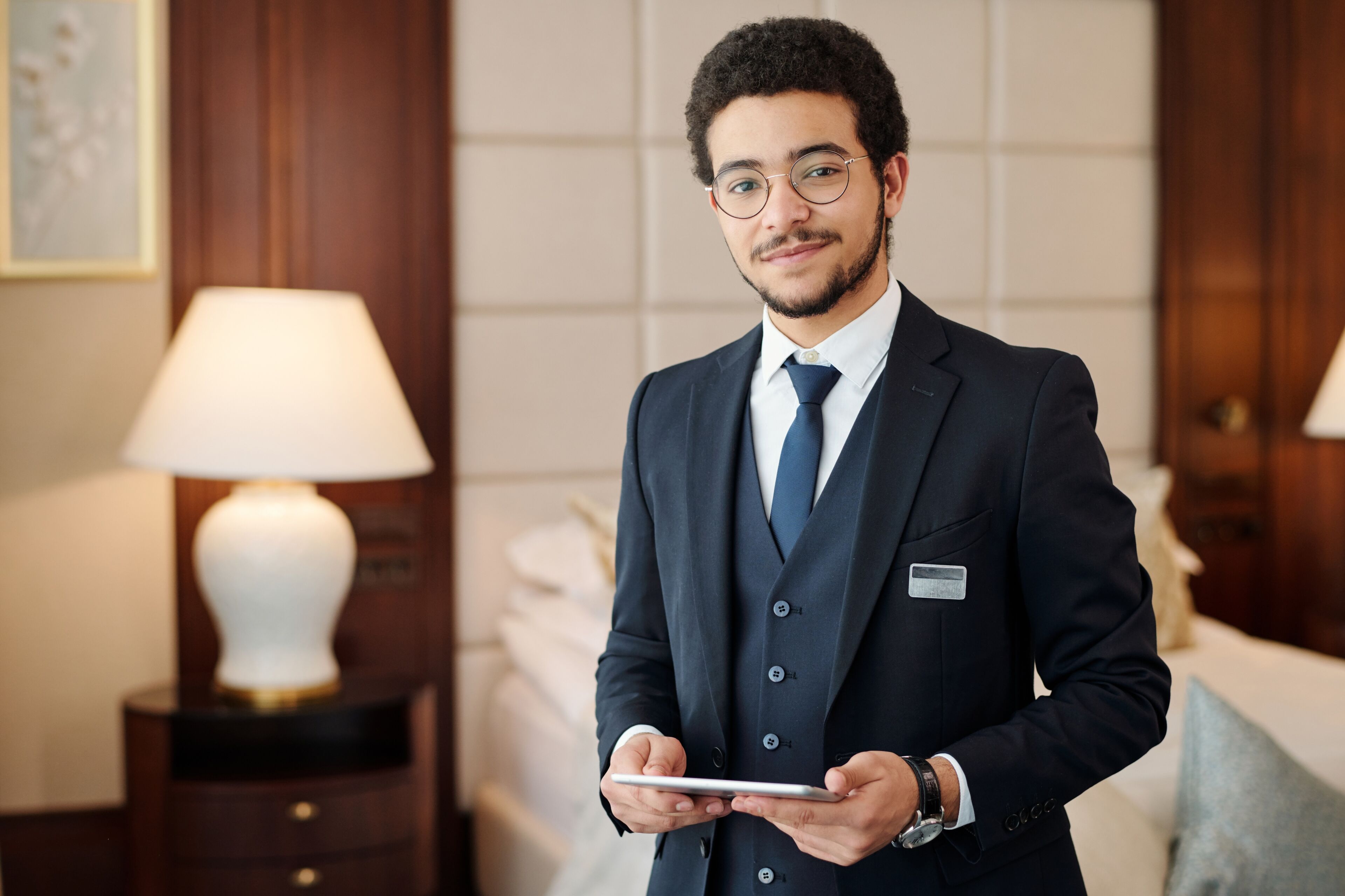 A professional male concierge in a tailored suit stands with a welcoming smile, holding a digital tablet in a luxurious hotel room.