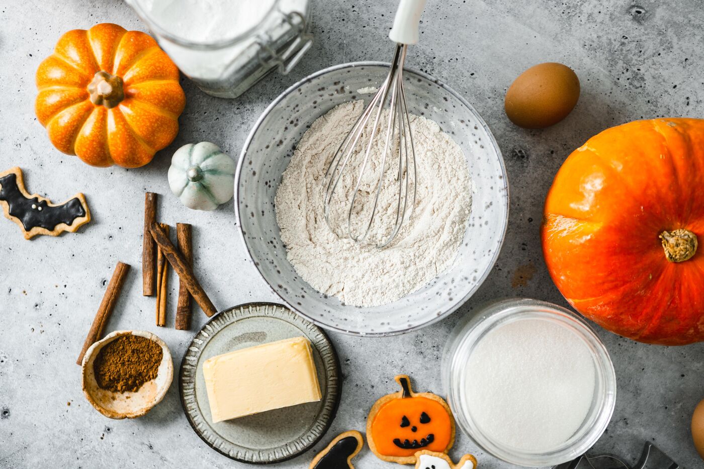 Ingredients for Halloween baking, including pumpkins, flour, eggs, butter, sugar, cinnamon, and spooky-shaped cookies, are laid out on a gray surface.
