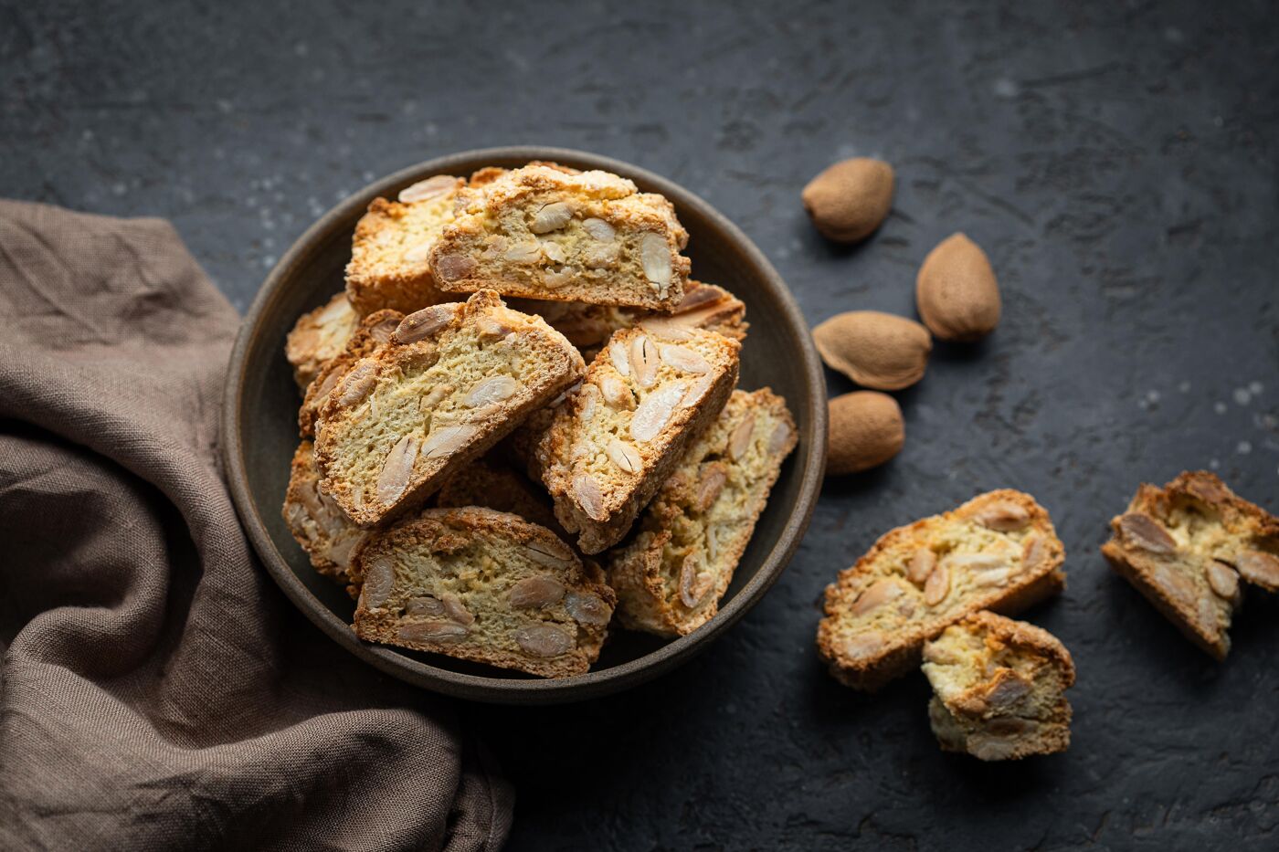 A bowl filled with crunchy, golden almond biscotti, with whole almonds scattered on a dark surface. These are an Italian classic, perfect for coffee or sweet wine.