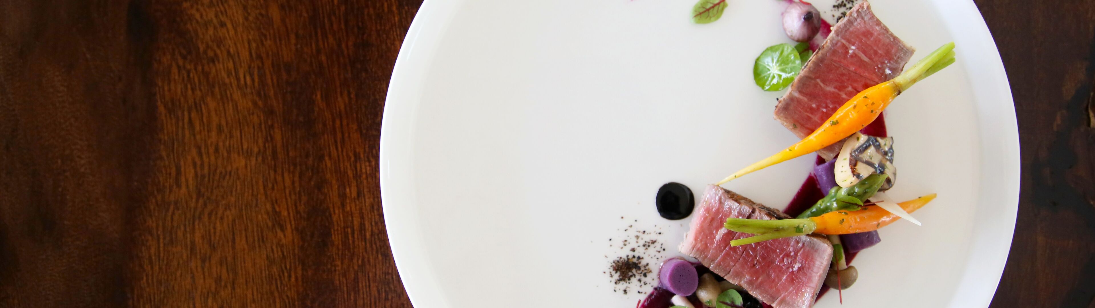 Présentation artistique de thon poêlé avec garnitures de légumes colorés sur une assiette blanche immaculée, sur une table en bois foncé.
