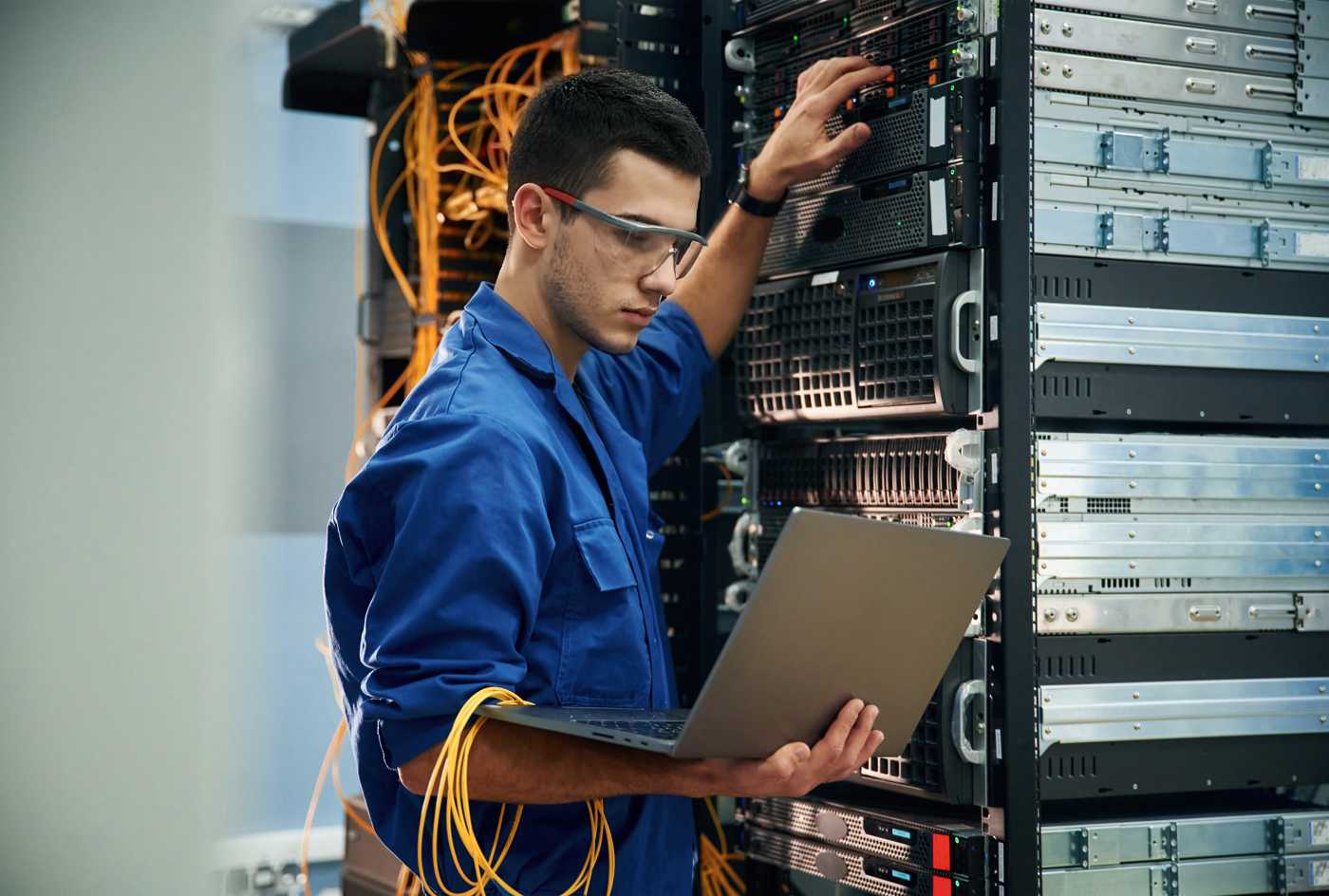 A technician wearing safety glasses and a blue jumpsuit works in a data center, handling cables and using a laptop to monitor servers.