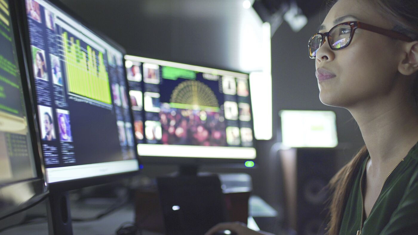 A focused professional woman working at a multi-monitor workstation, analyzing data.