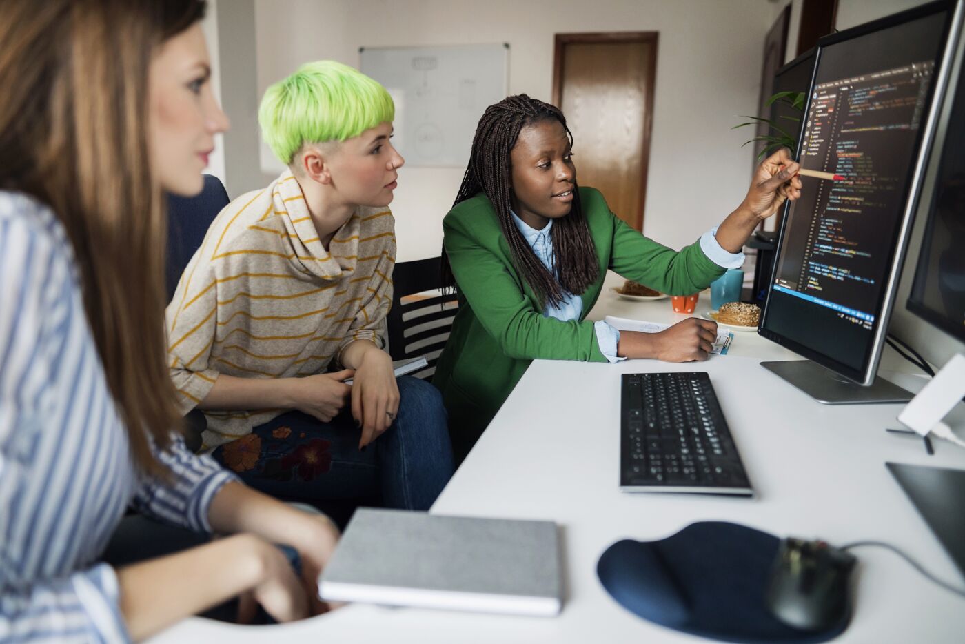 Three individuals collaborating on a coding project, one pointing at a computer screen with software code.