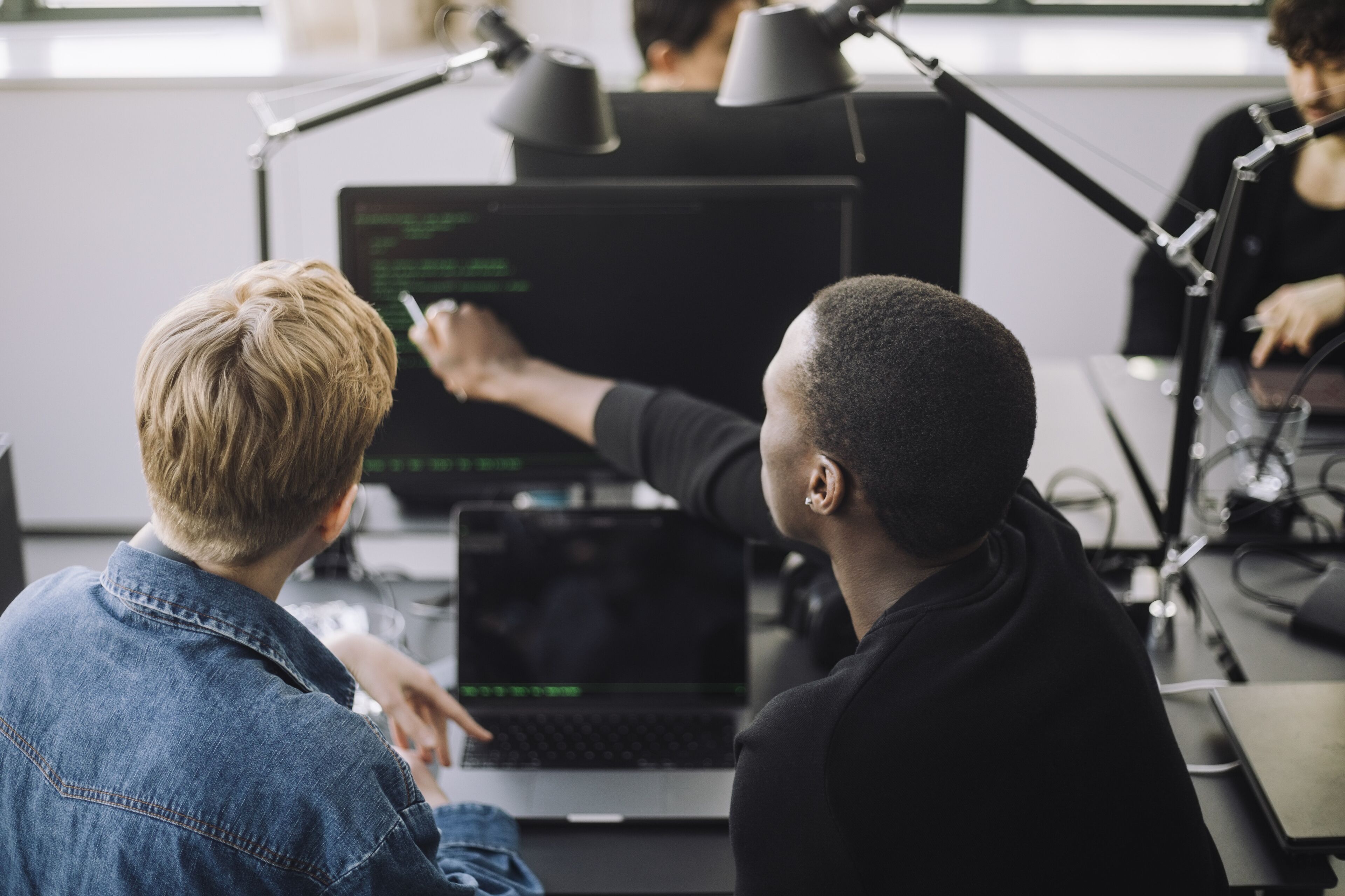 Dos individus en una tasca de programació col·laborativa, un assenyalant una pantalla d'ordinador en un entorn d'oficina.