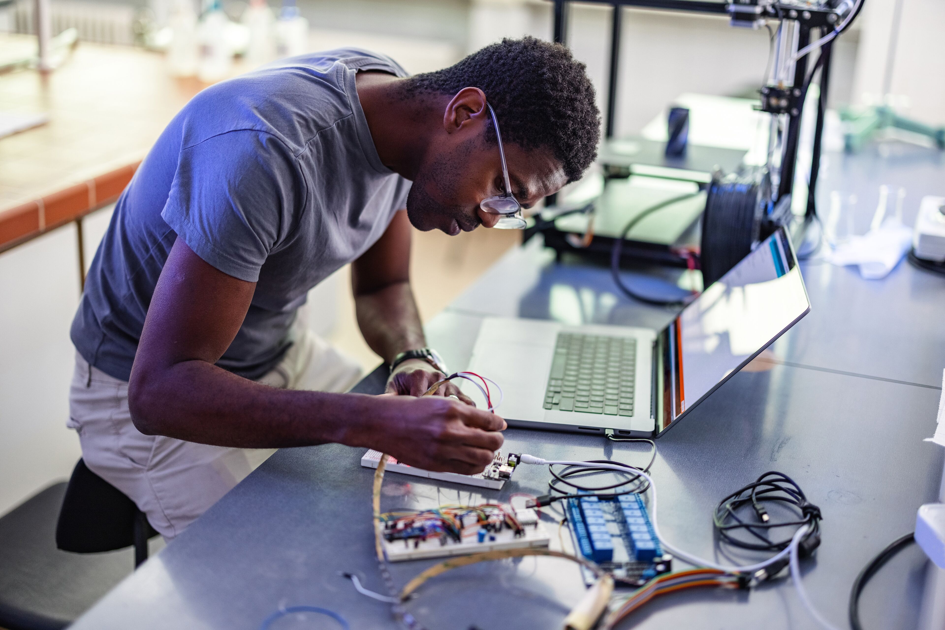 Una persona concentrada està soldant una placa de circuit en un espai de treball ple de tecnologia, amb un portàtil a prop.