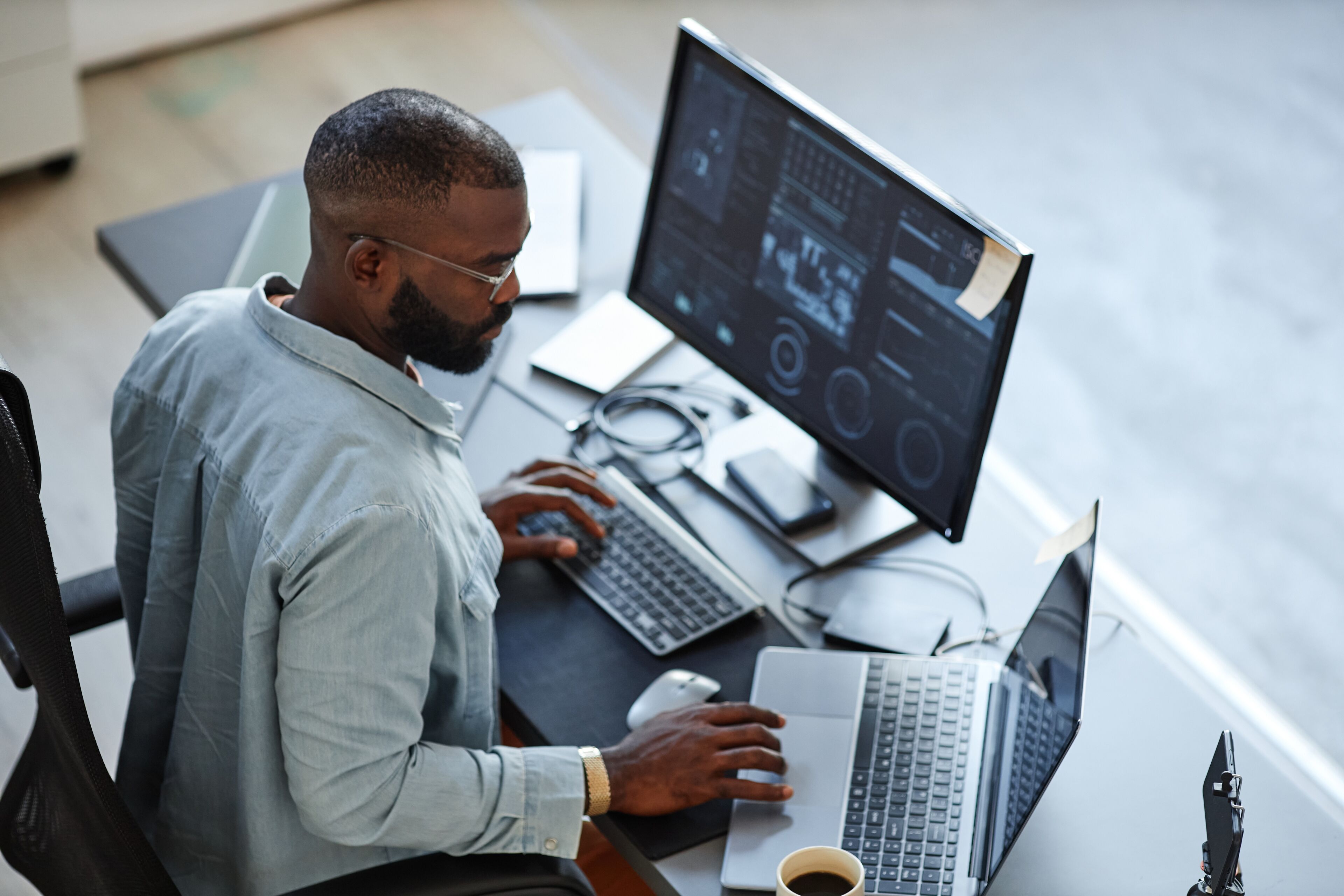 Un hombre enfocado trabaja en una configuración de múltiples monitores, rodeado de gadgets tecnológicos, reflejando un espacio de trabajo profesional y moderno.