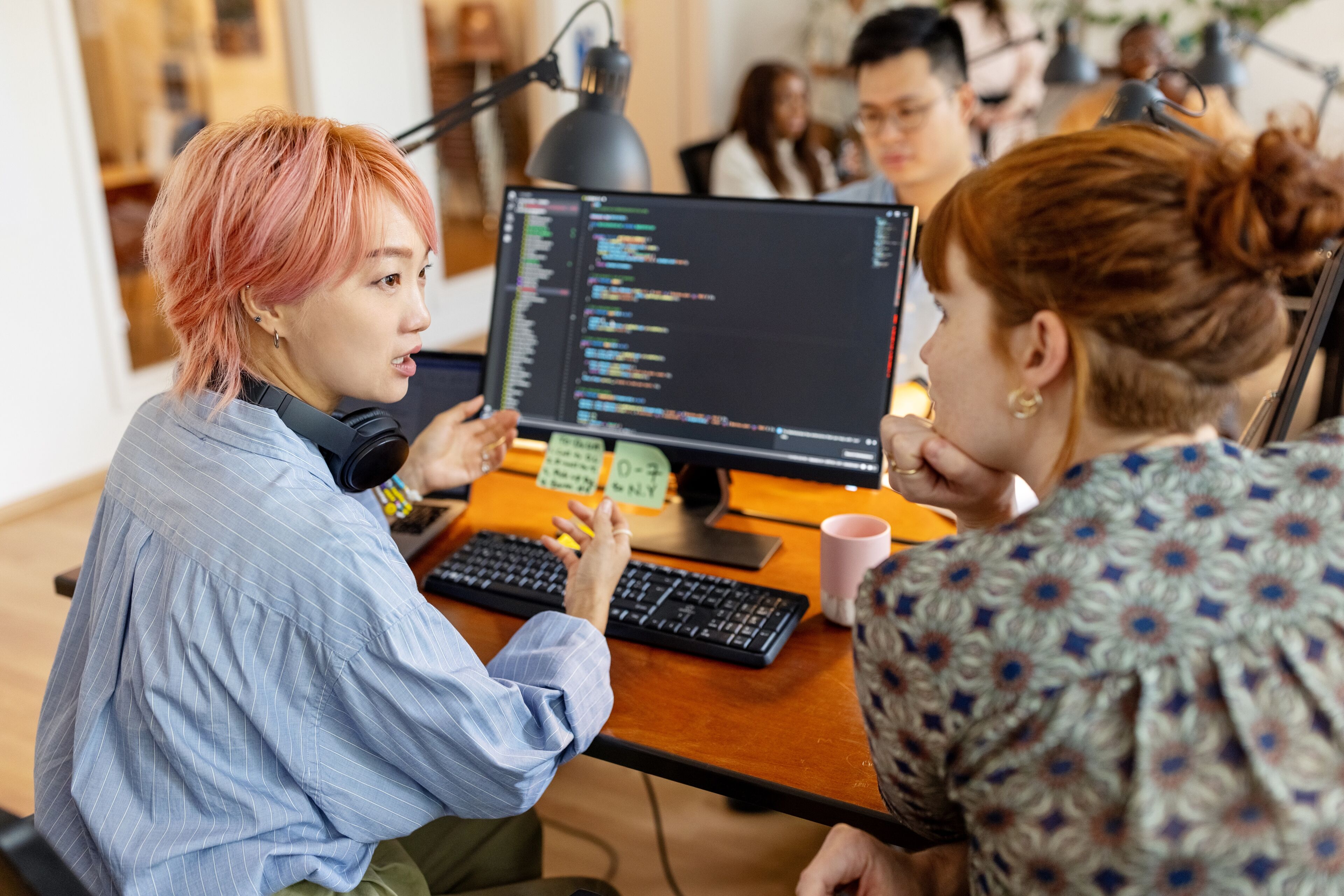 Una desarrolladora con cabello rosa hablando de código con un colega en una oficina concurrida.
