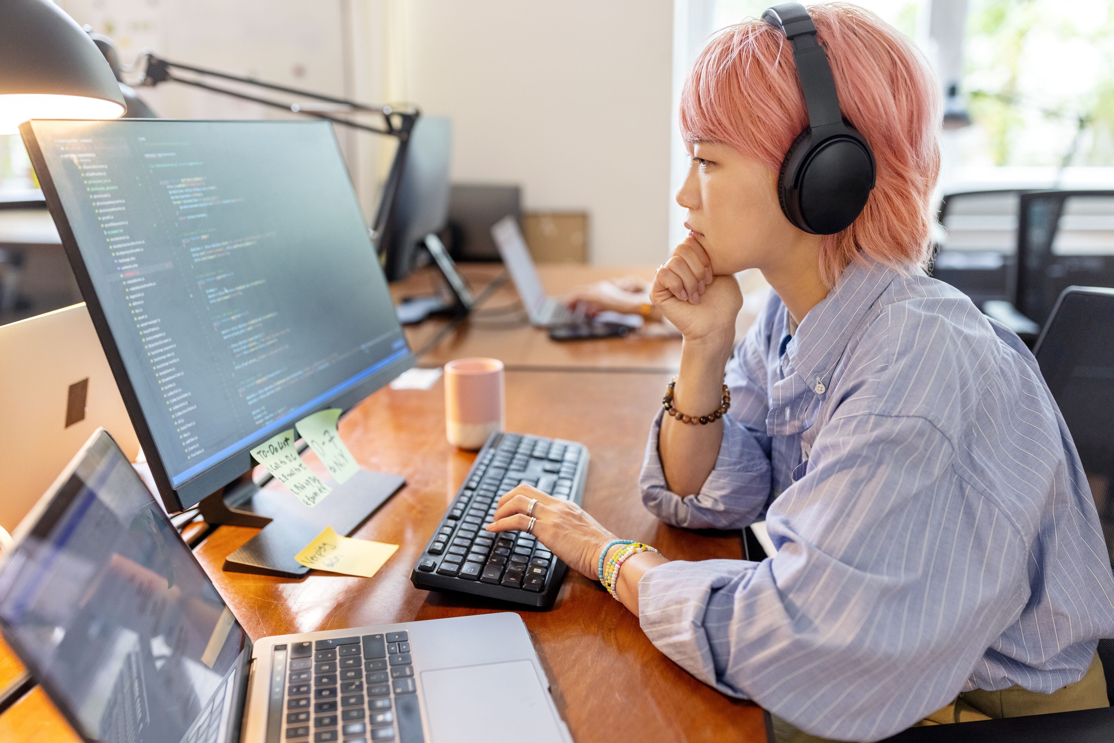 A focused individual with pink hair, wearing headphones, codes on a computer in a well-lit office.