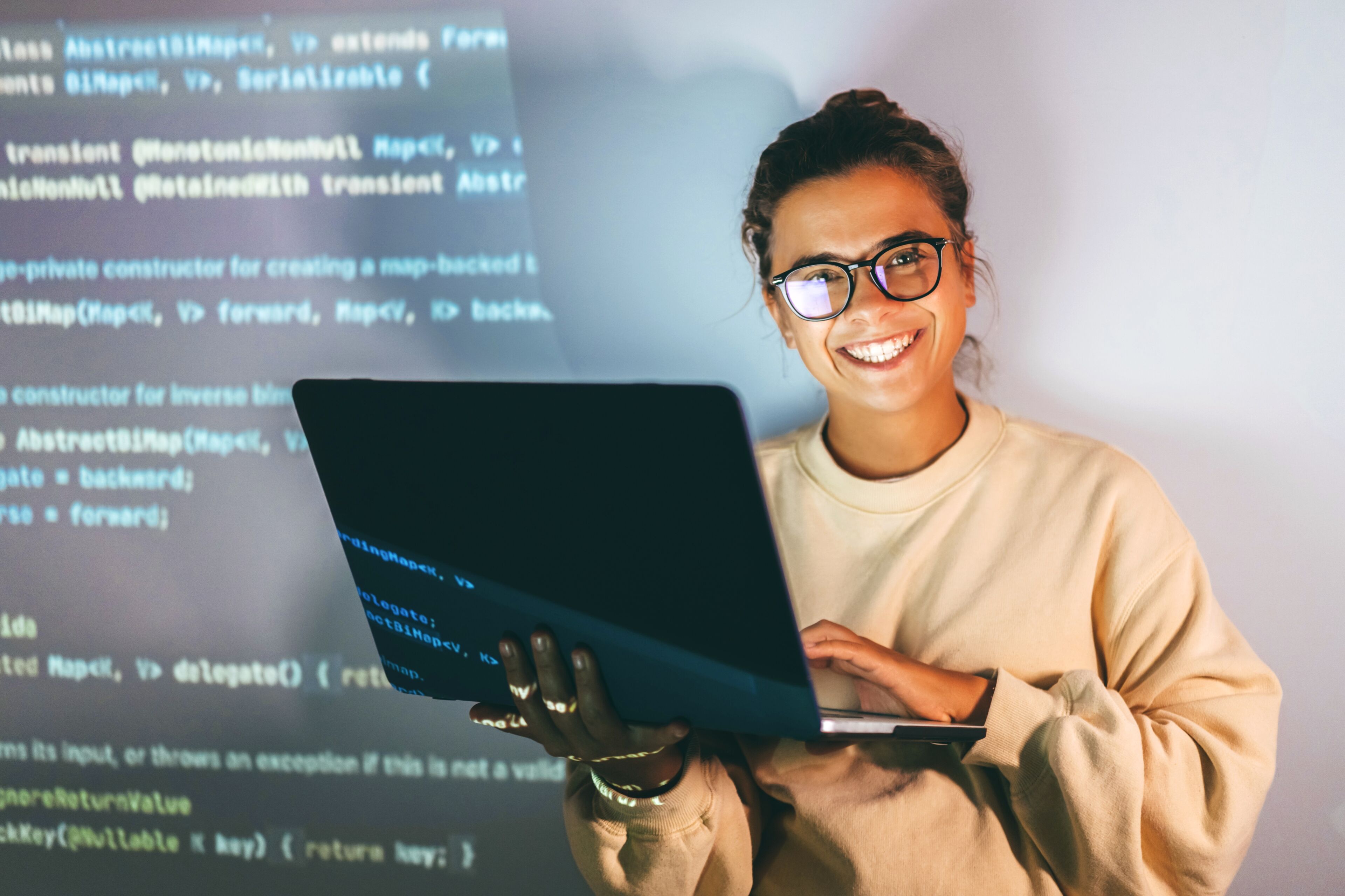 Une femme souriante aux lunettes tenant un ordinateur affichant du code, dégageant un sentiment de réussite et d'expertise en programmation.