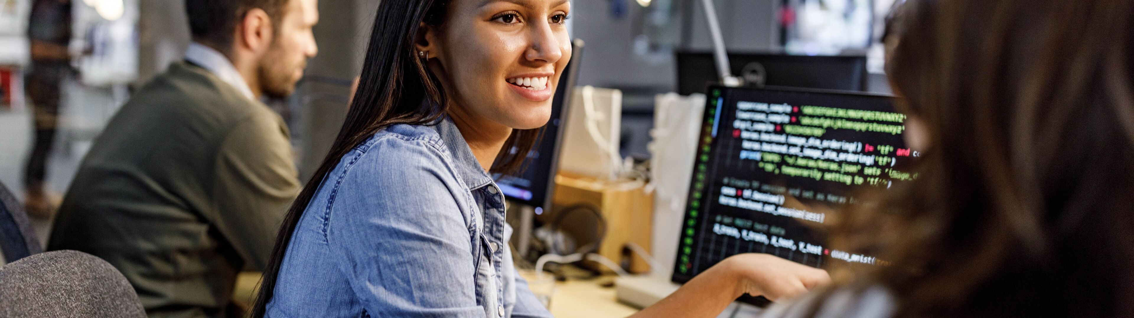 Une développeuse souriante programme sur un ordinateur, collègues en arrière-plan.