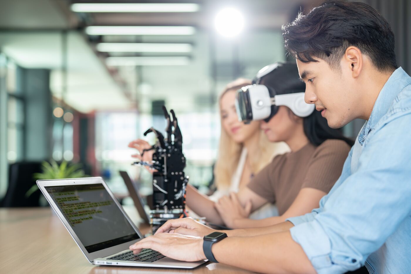 A diverse tech team collaborates in a modern workspace; one member uses a VR headset, another interacts with a robotic arm, while a third focuses on coding on a laptop.