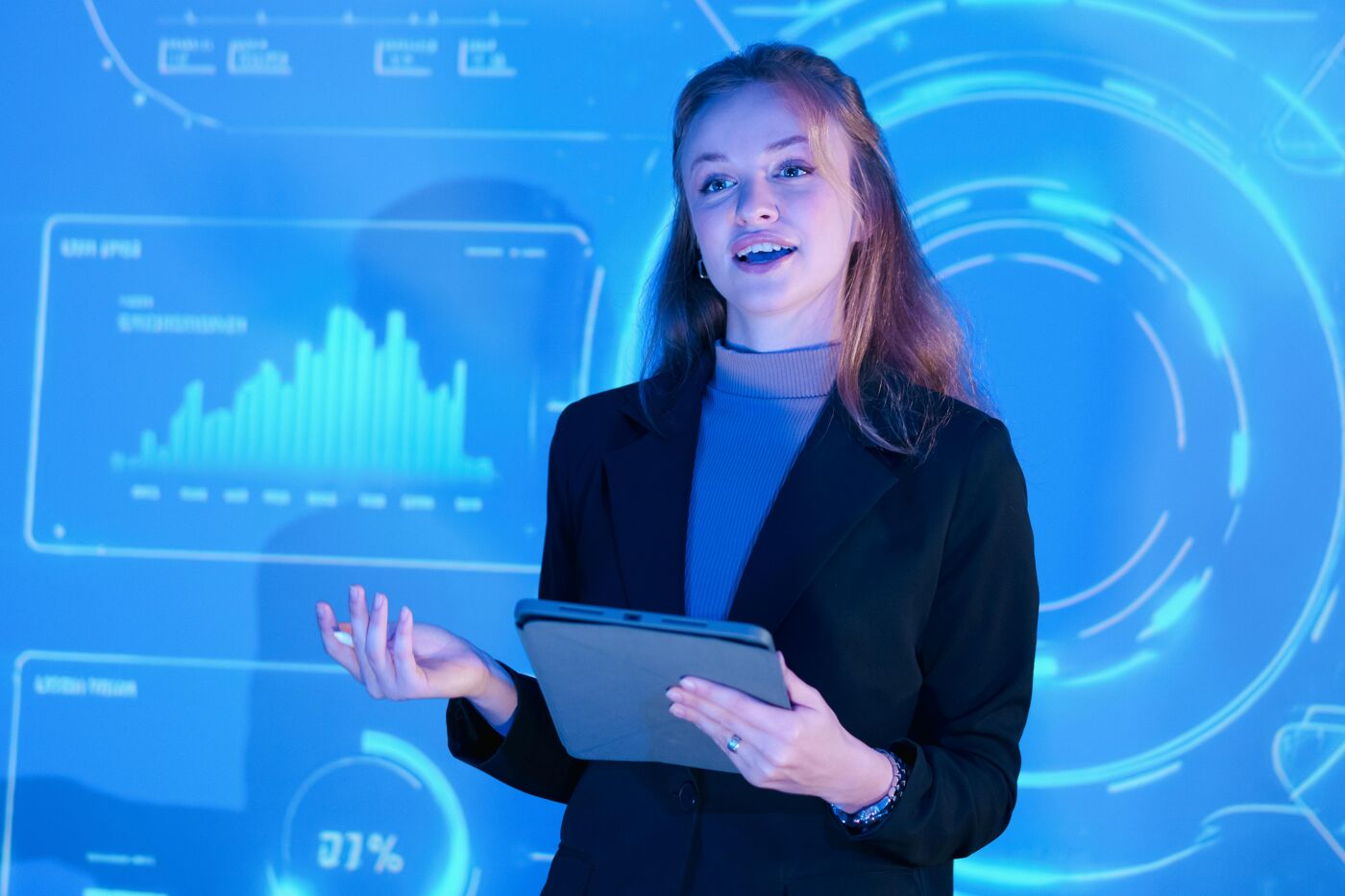 A young woman in a dark jacket is giving a presentation in front of a blue screen displaying charts and circles. She is holding a tablet.