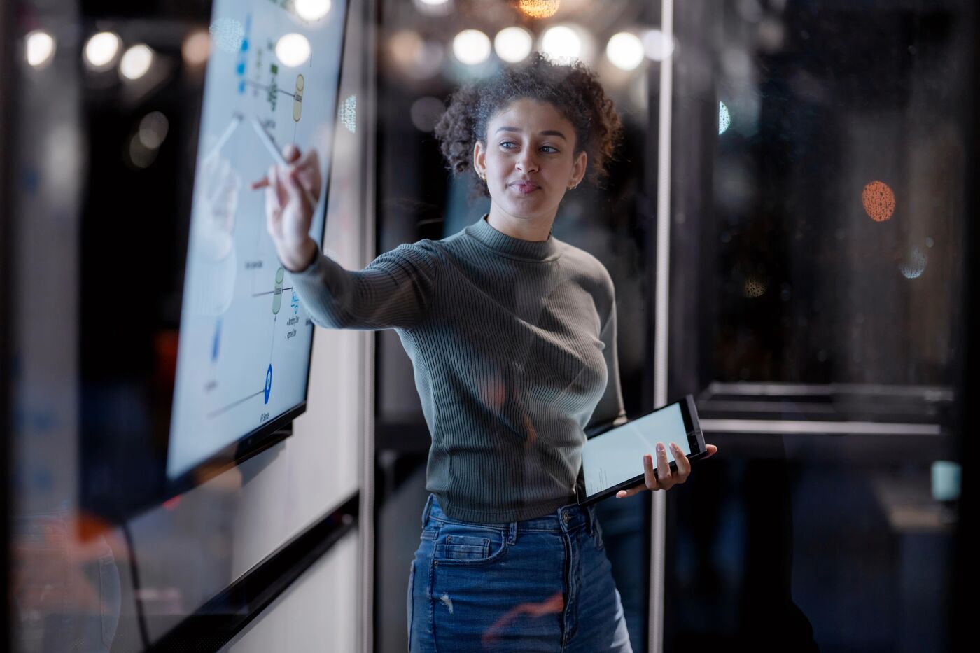 A young woman with curly hair is pointing at a digital screen displaying charts, while holding a tablet in her other hand.