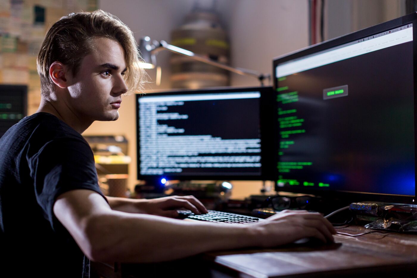 A young man with blond hair styled back is seated at a computer, typing on a keyboard. He is focused on the screens displaying code.