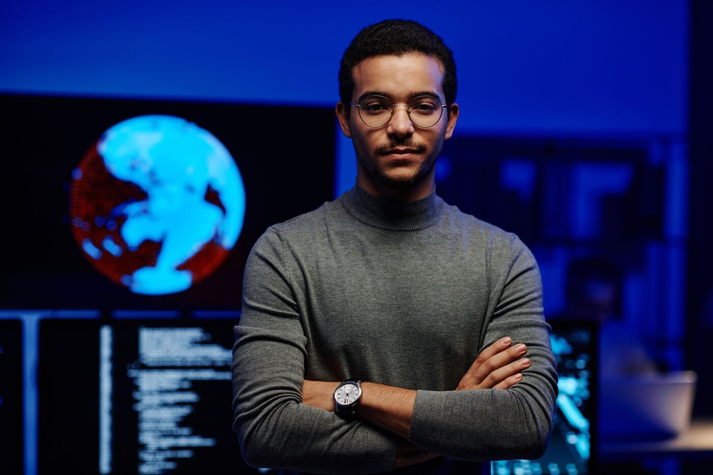 Confident young professional with glasses, standing arms crossed in a high-tech workspace, with a glowing digital globe in the background.