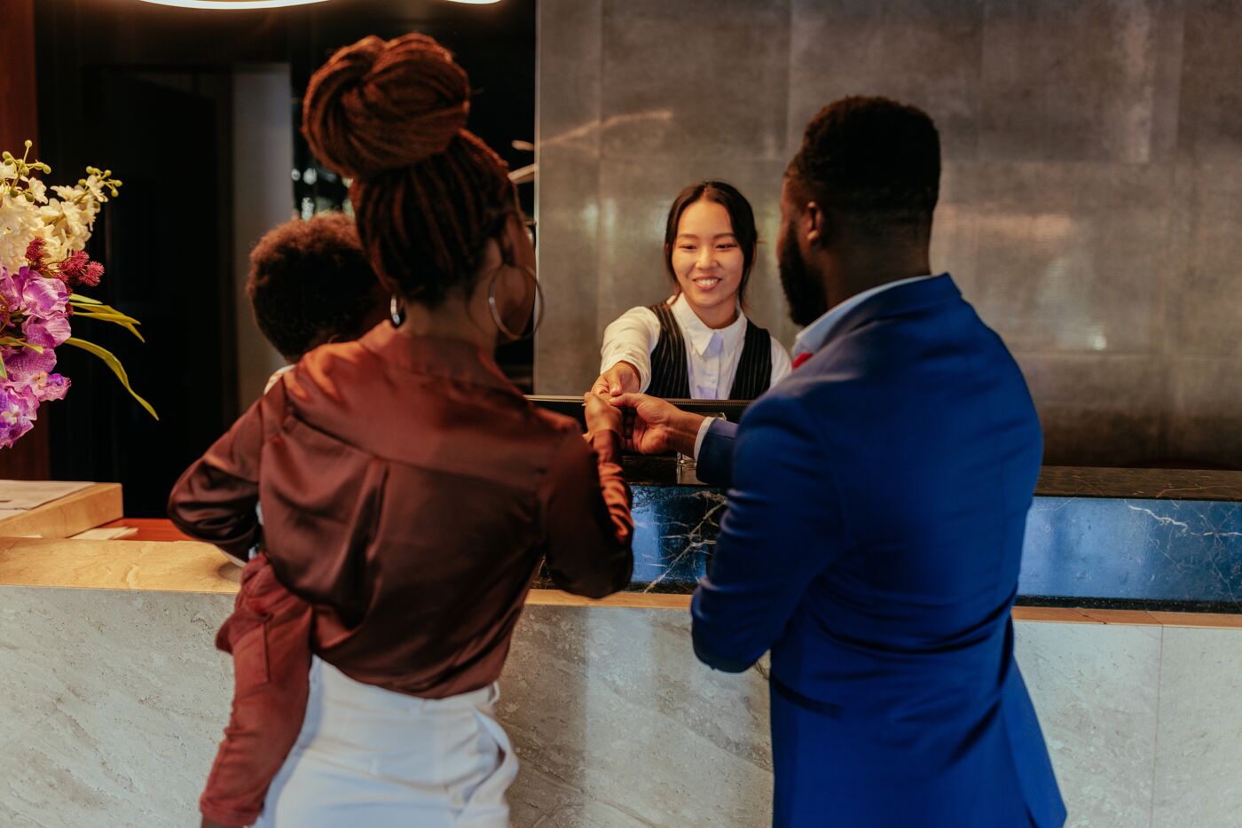 A man in a blue suit and a woman with an updo hairstyle are checking in at a hotel reception, greeted warmly by the smiling receptionist.