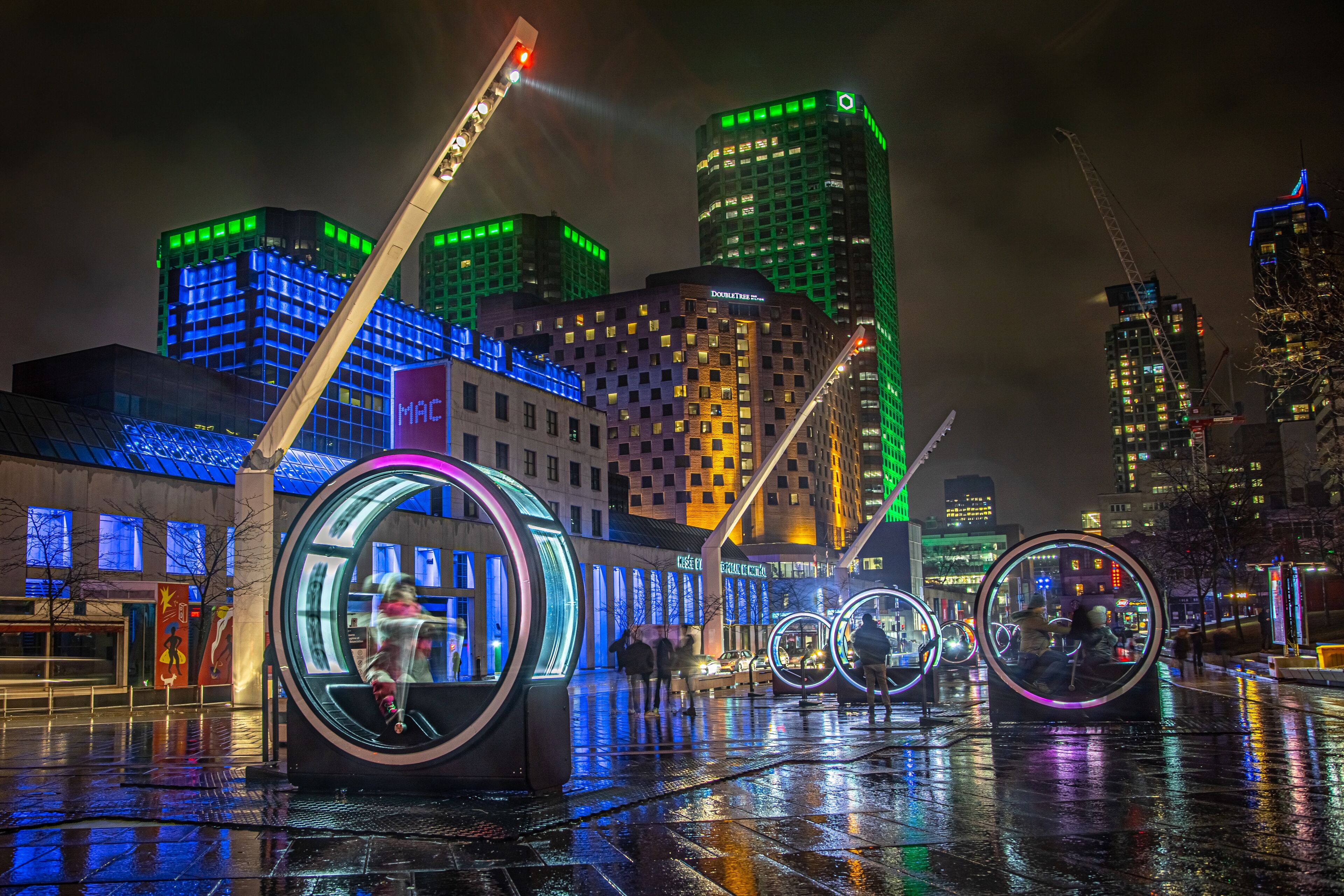 A vibrant cityscape at night featuring futuristic light installations with illuminated circular pods that people can walk through, set against a backdrop of brightly lit skyscrapers.