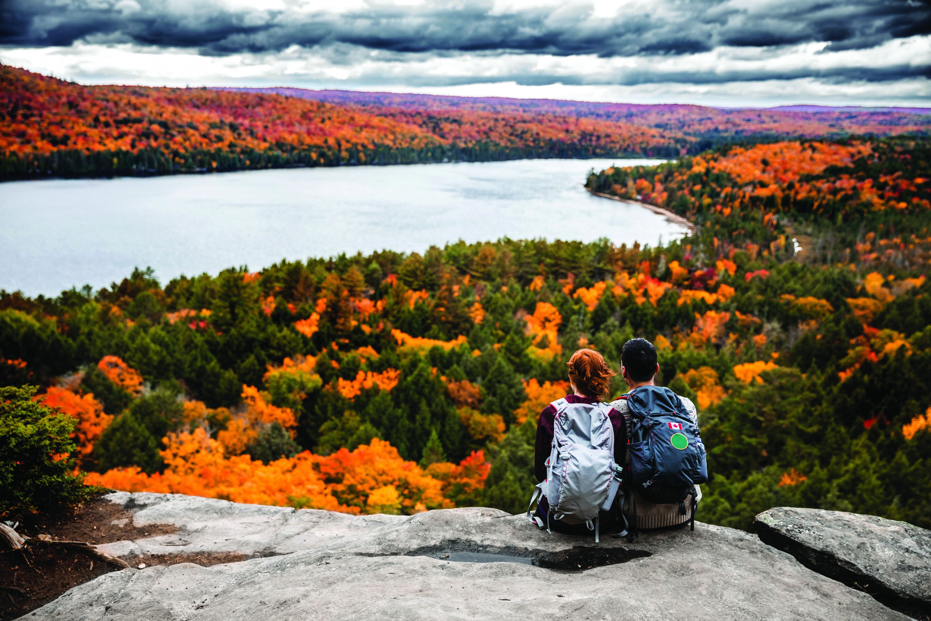 Two hikers sit on a cliff overlooking a stunning autumn landscape filled with fiery foliage by a serene lake, capturing the essence of fall exploration.