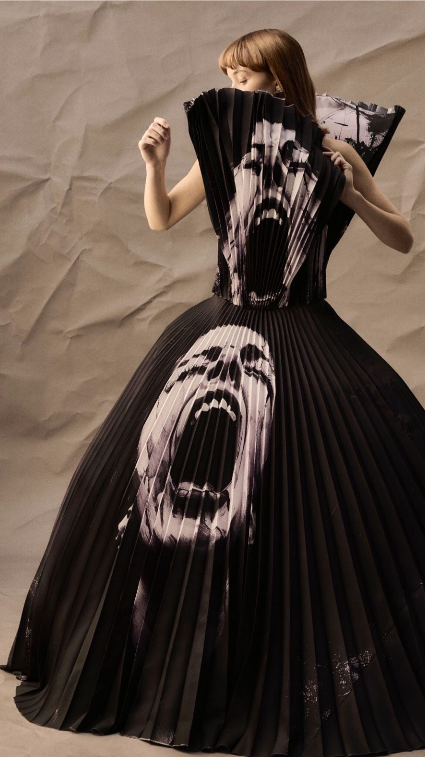 A black gown featuring a screaming face design in black and white, with a pleated collar and skirt. The image has a crumpled paper background.